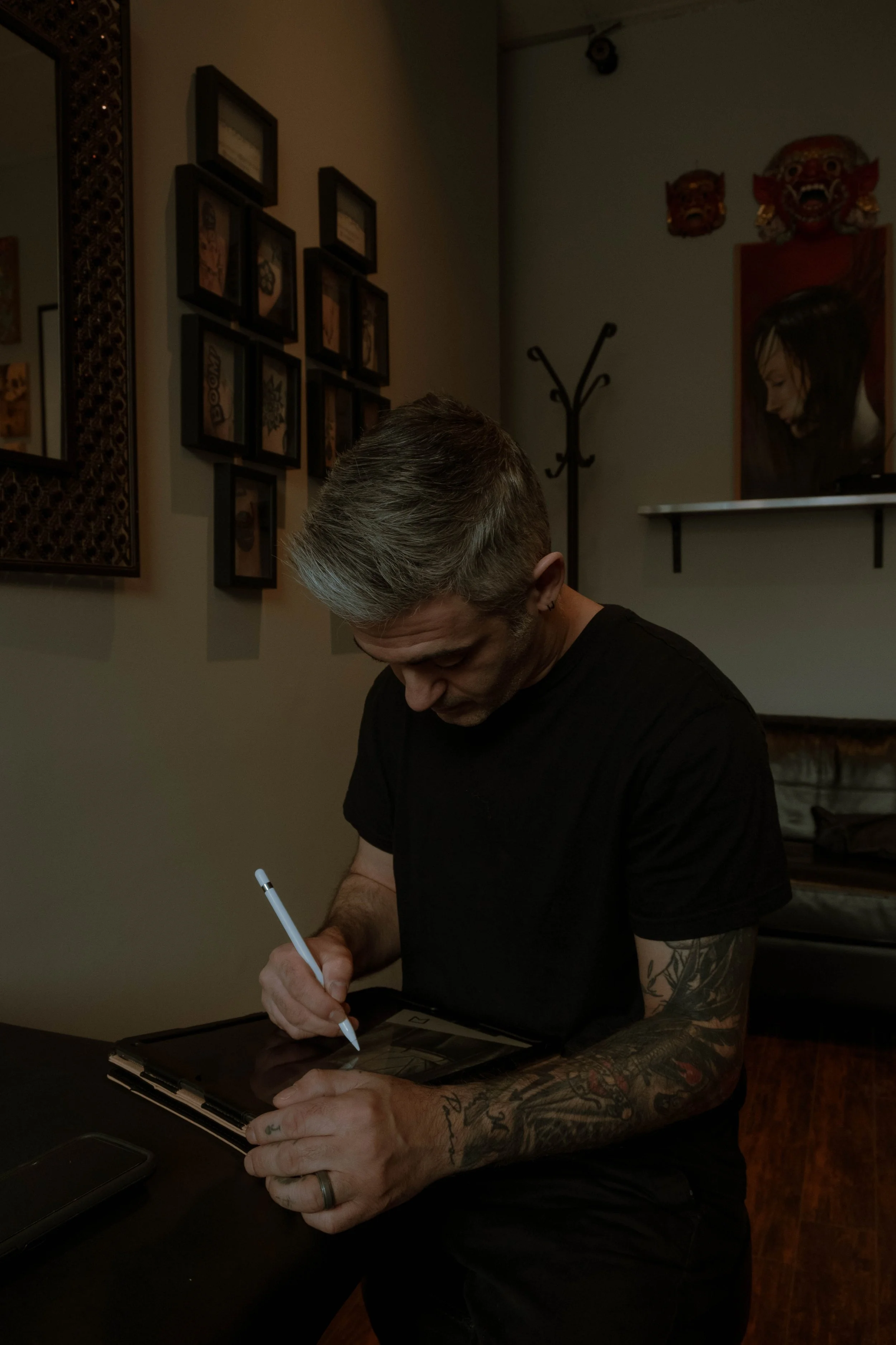 A man with gray and black hair, tattoos on his arms, wearing a black t-shirt, is sitting at a table signing a photo or piece of artwork with a white pen. The background shows a wall with artwork, masks, and a coat rack.