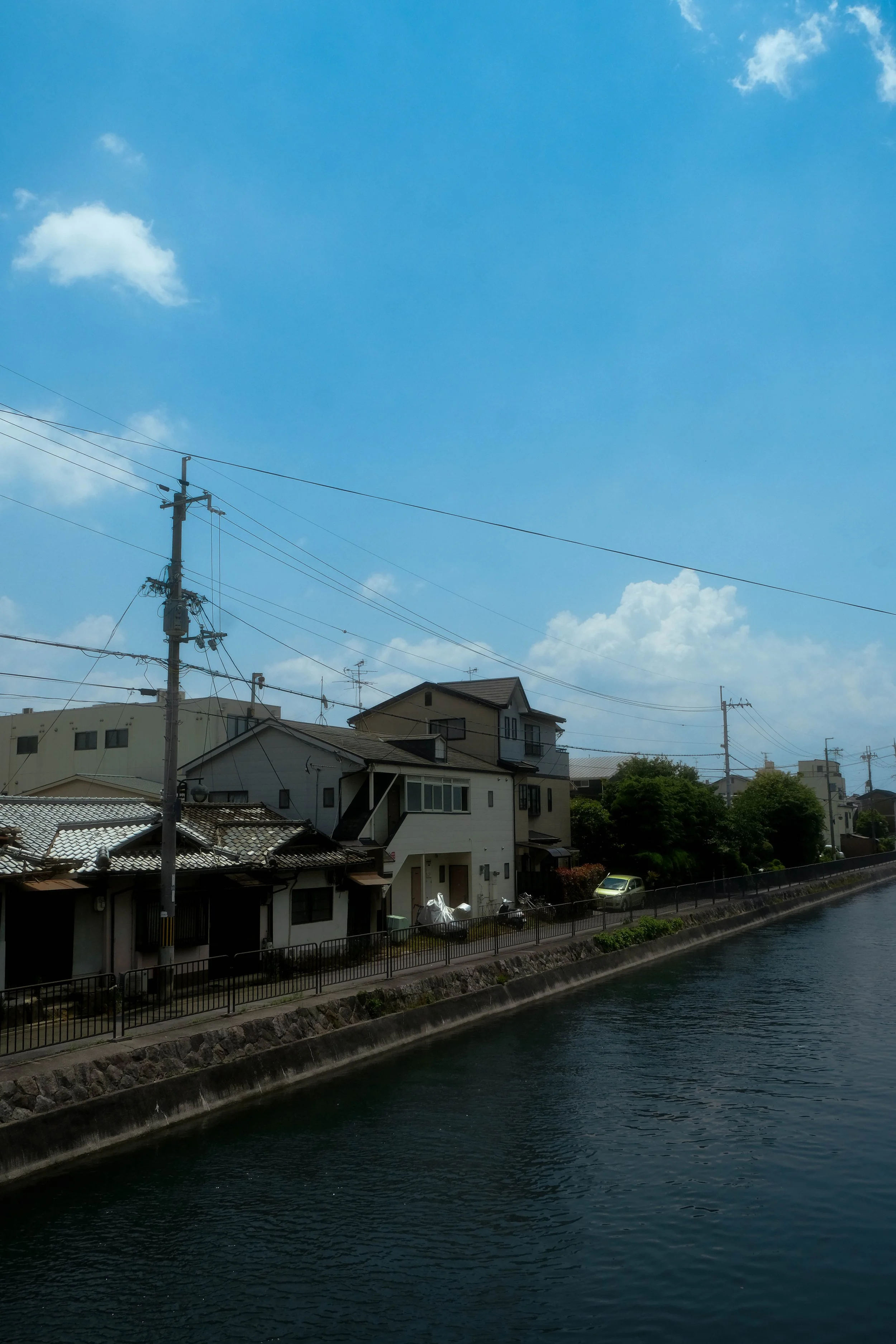 Residential houses along a canal with a stone embankment, power lines, and a bright blue sky with a few clouds.