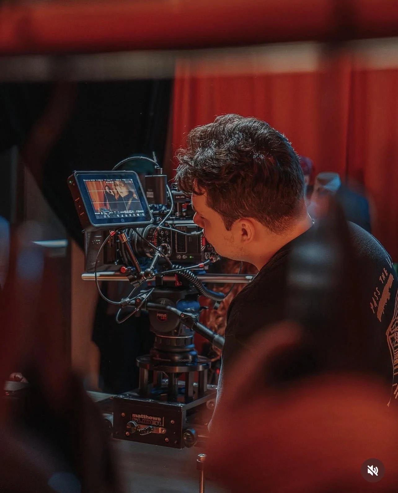 A film director or camera operator adjusting a professional camera on a film set with red curtains in the background