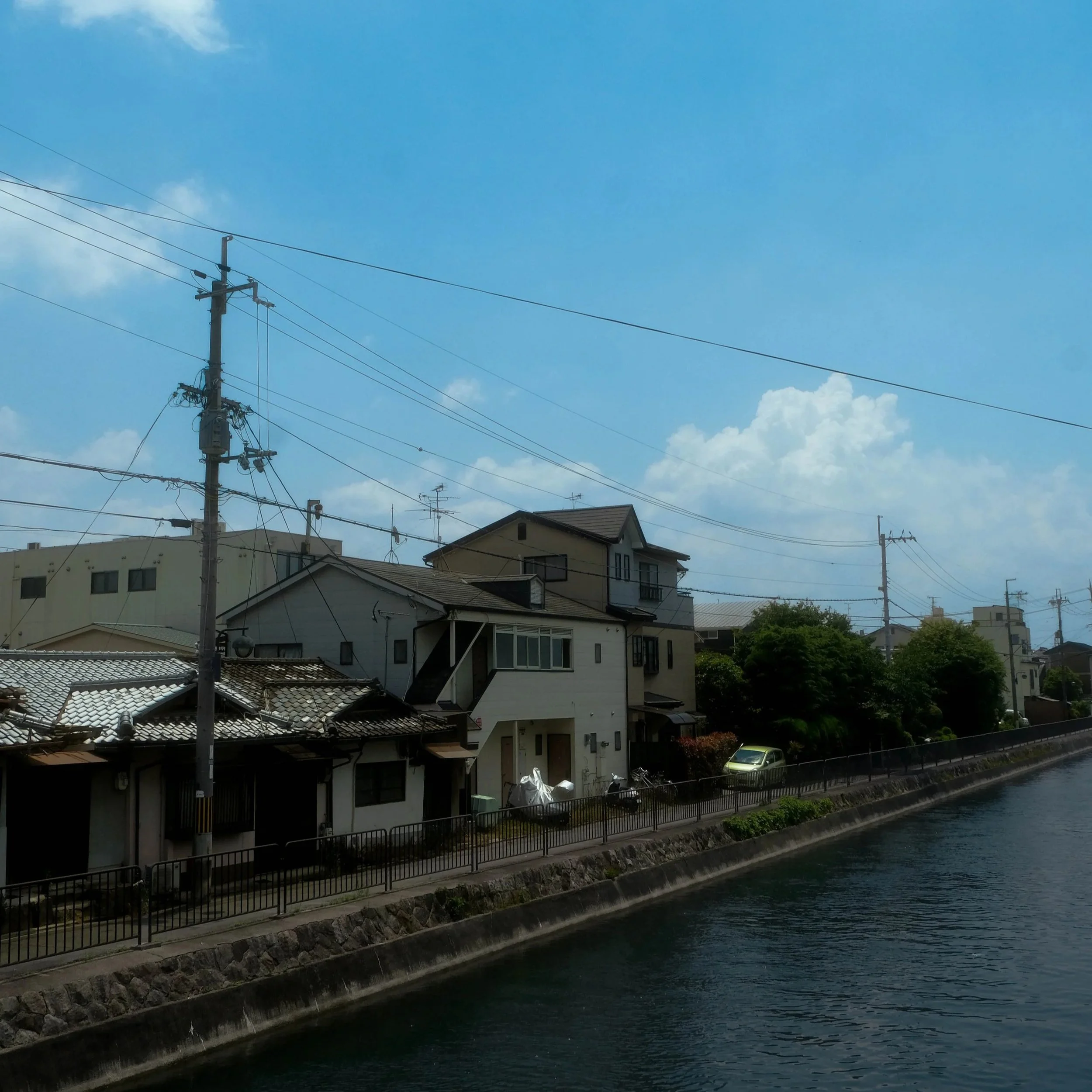 Residential houses along a canal with a waterway at the bottom, and power lines with a utility pole in the foreground, under a mostly clear blue sky.
