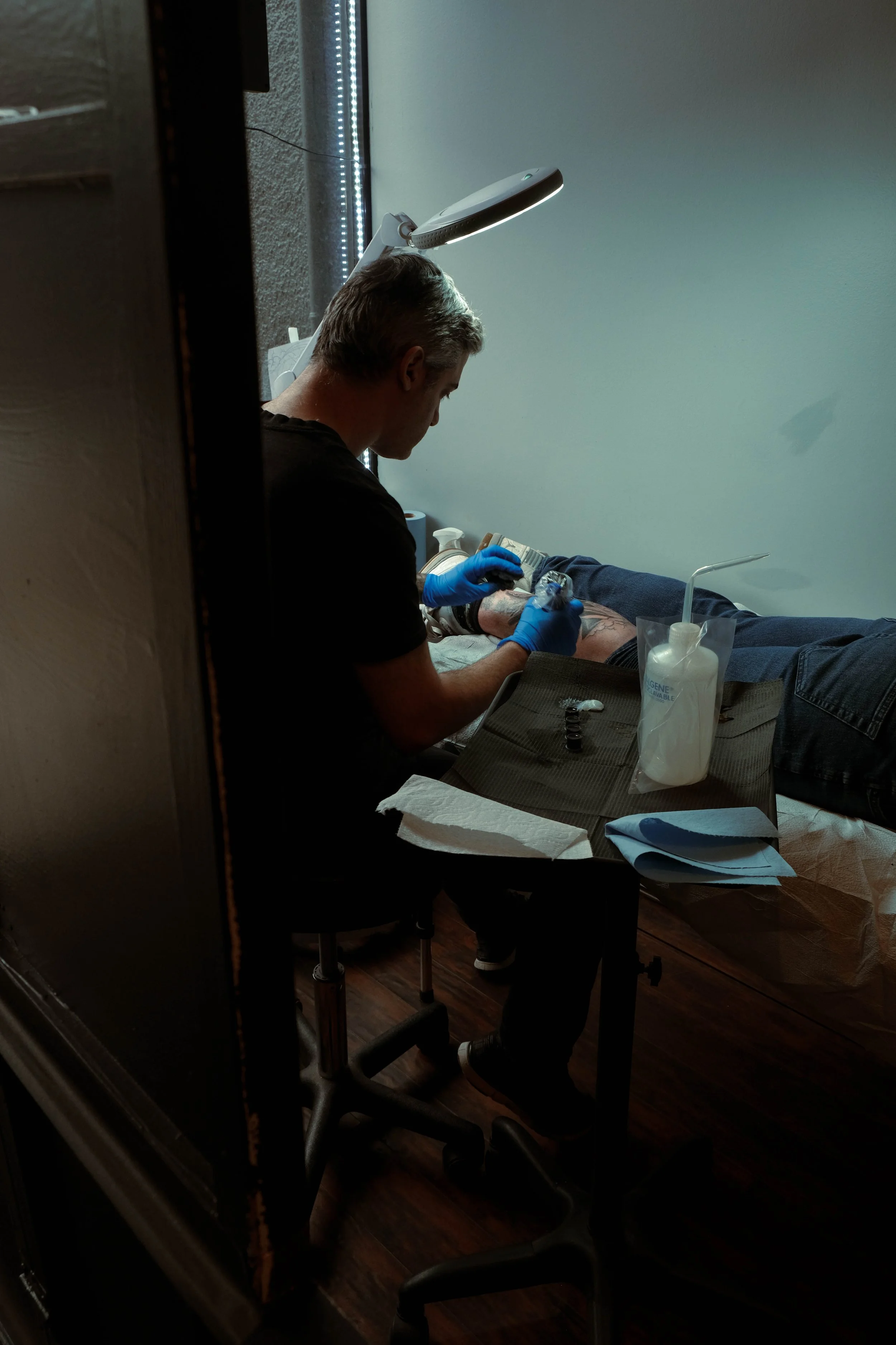 A tattoo artist wearing blue gloves is working on a client's leg in a small room with a white wall. The artist is focused on creating a tattoo, and there are tattoo supplies on the table, including ink caps, paper towels, and lotion. A large adjustable lamp is illuminating the work area.