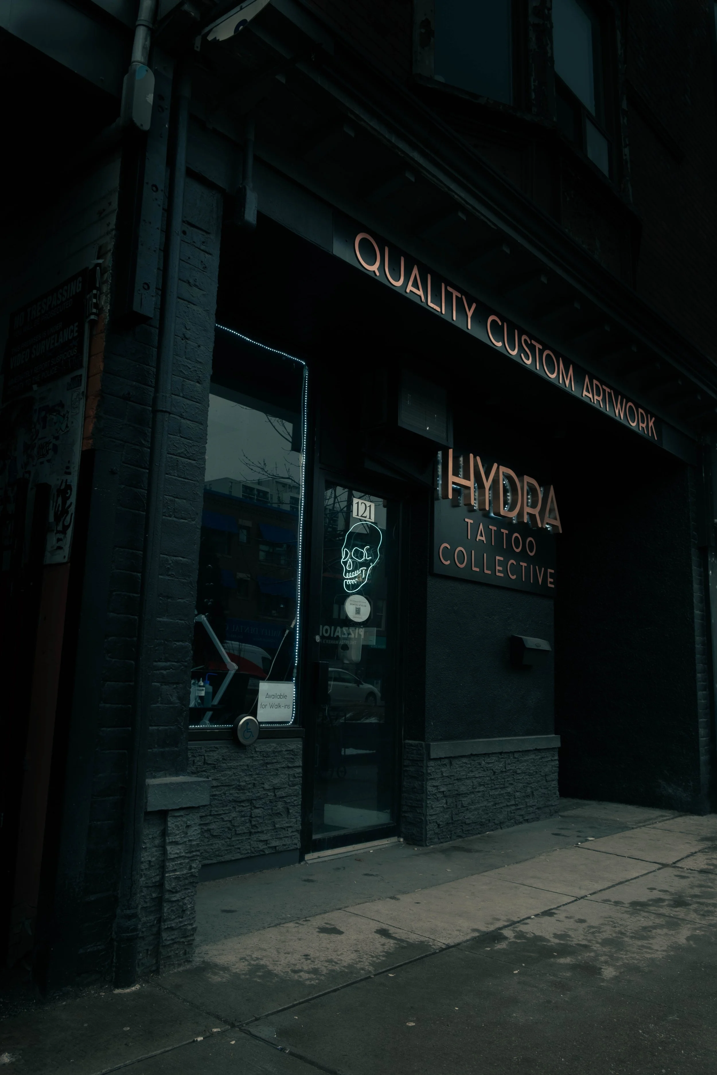 storefront with the sign 'Quality Custom Artwork' and 'Hydra Tattoo Collective' on the building, window with a neon skull sign, and a sidewalk in front.