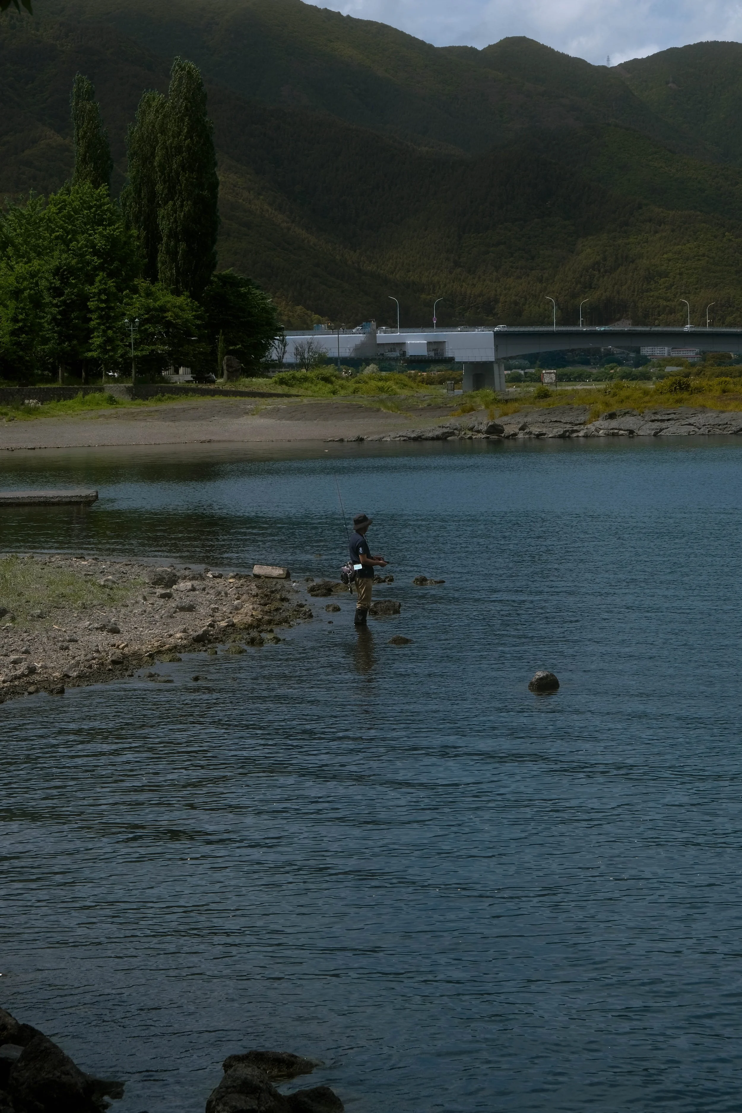 A person is fishing in a river with a mountainous landscape in the background. The sky is partly cloudy, and there is a bridge crossing over the river.