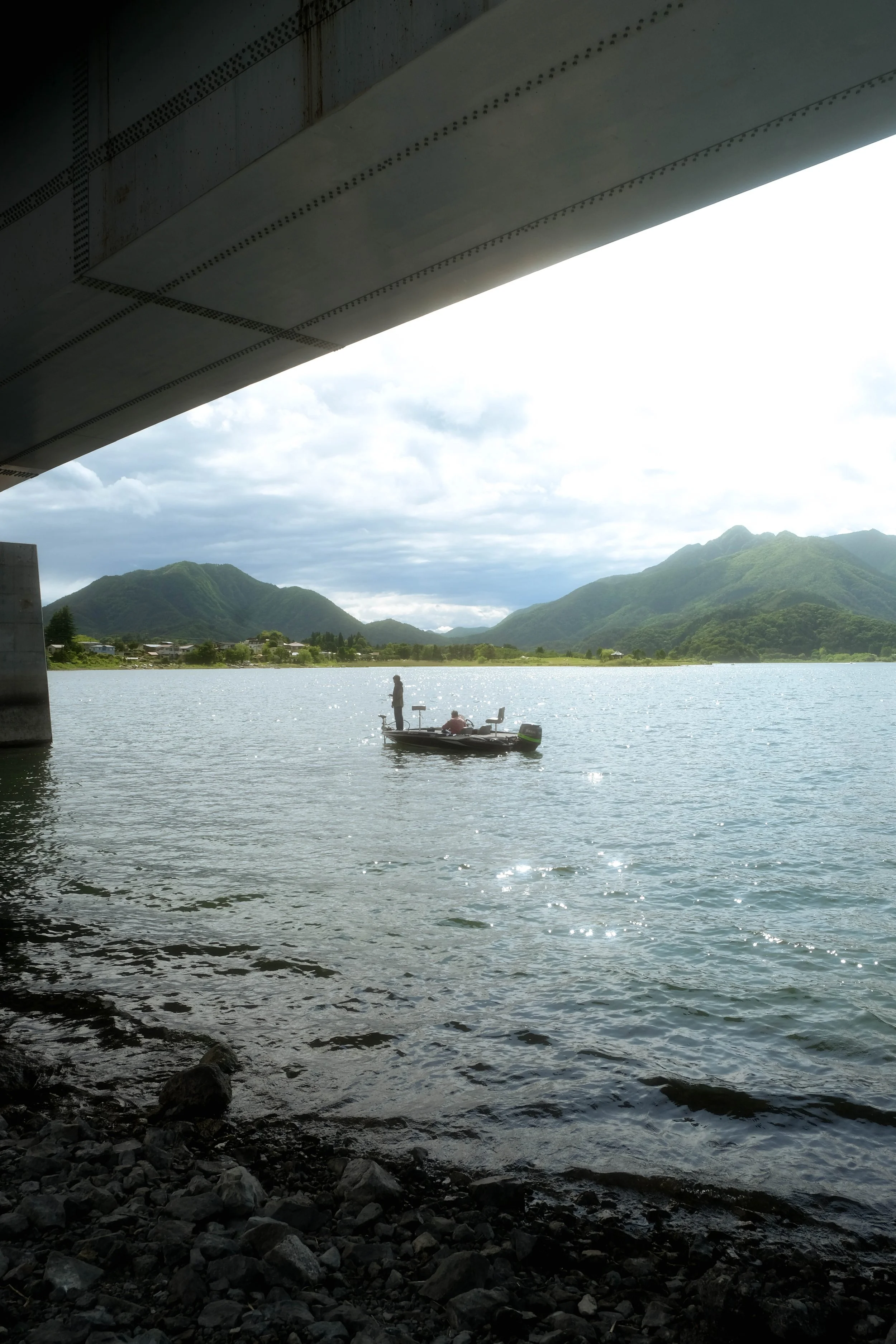 View of a river with mountains in the background, seen from under a bridge. There are two people on a small boat in the water.