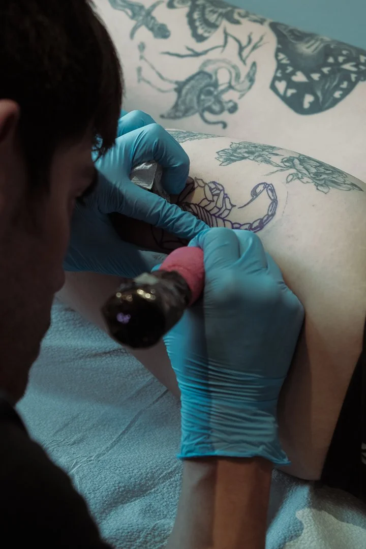 Tattoo artist wearing blue gloves sketches a tattoo design on a client's skin using a tattoo machine.