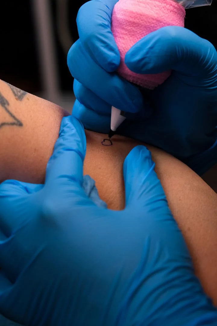 A tattoo artist wearing blue gloves is tattooing a small heart on someone's skin with a tattoo needle.