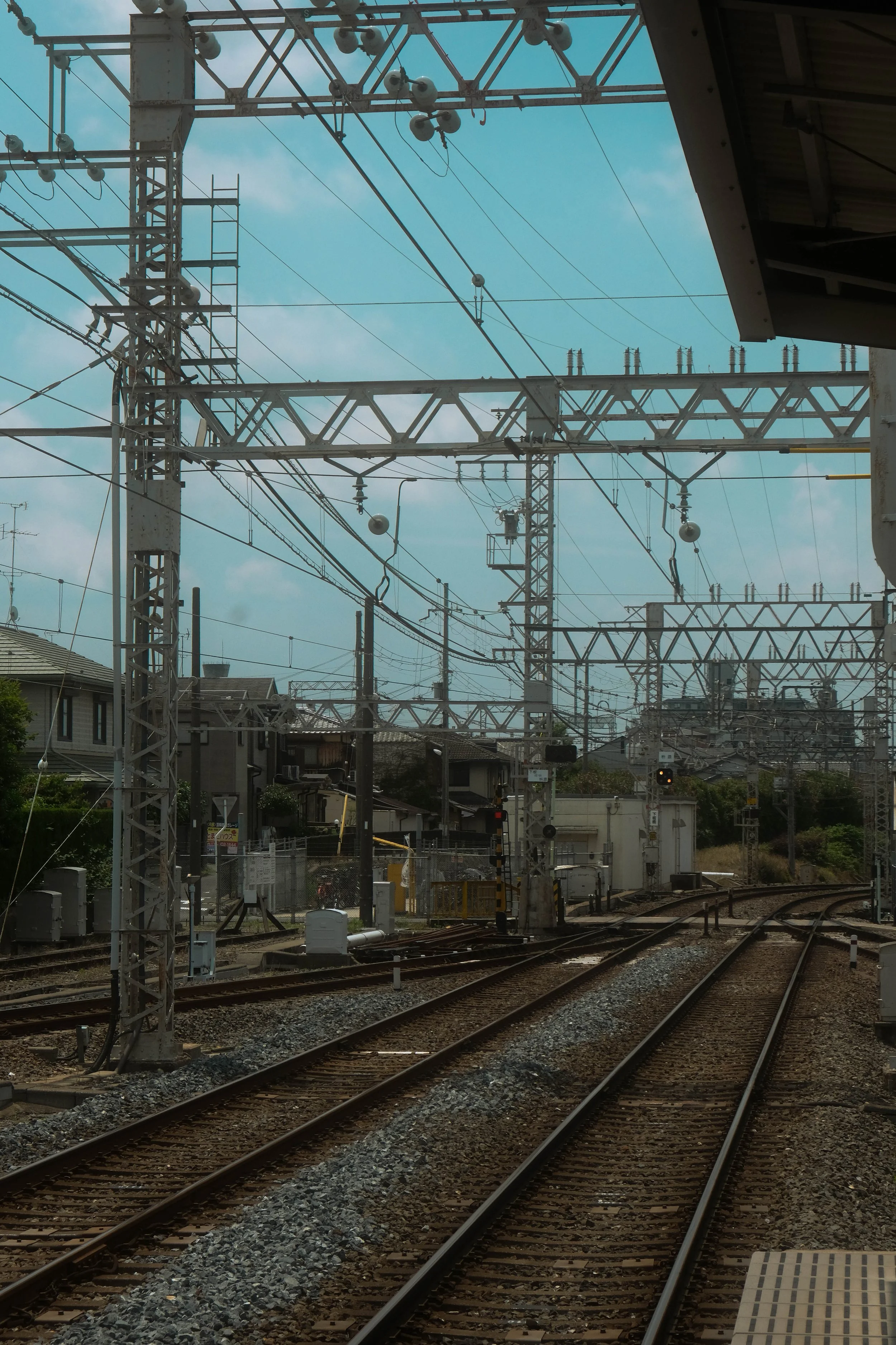View of railway tracks with overhead electrical wires and equipment, houses and trees in the background, under a partly cloudy sky.