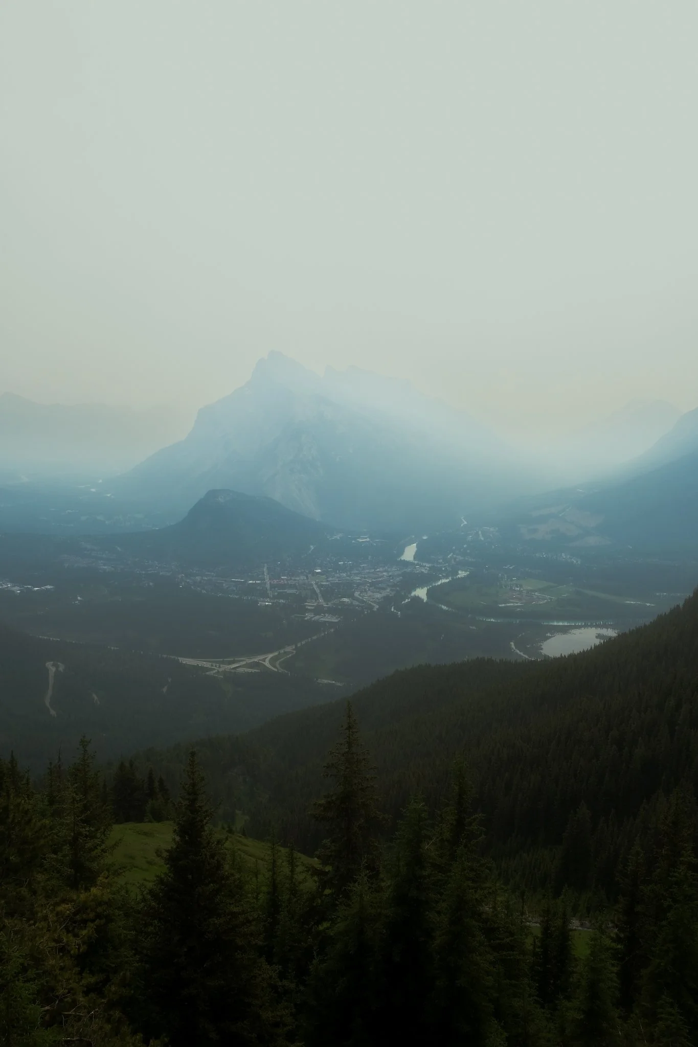 Mountain landscape with forested foreground, winding roads, and a river, slightly obscured by mist or haze.