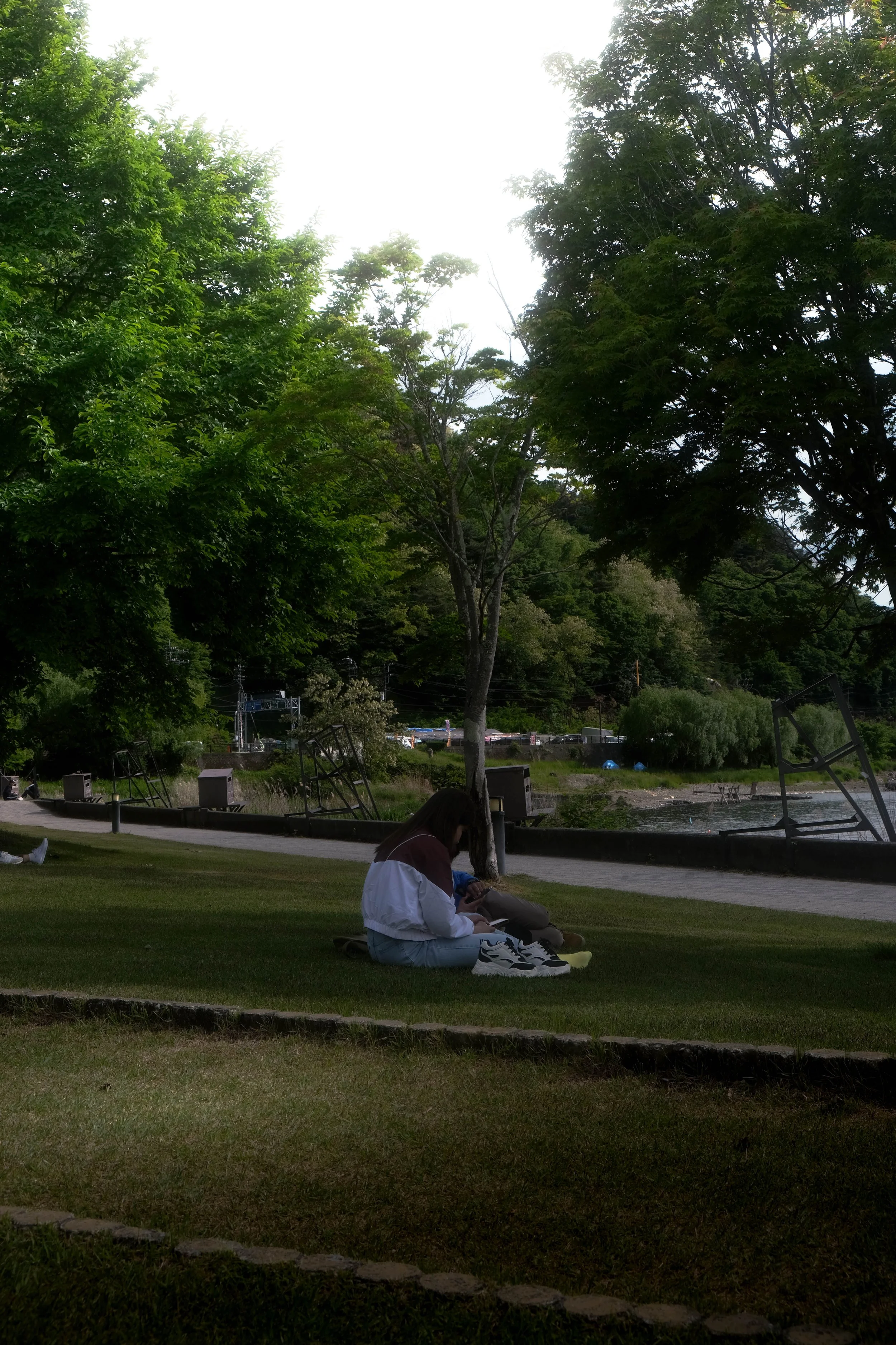 A person sitting on a grassy area near a river, with their shoes off beside them, surrounded by trees in a park during daytime.