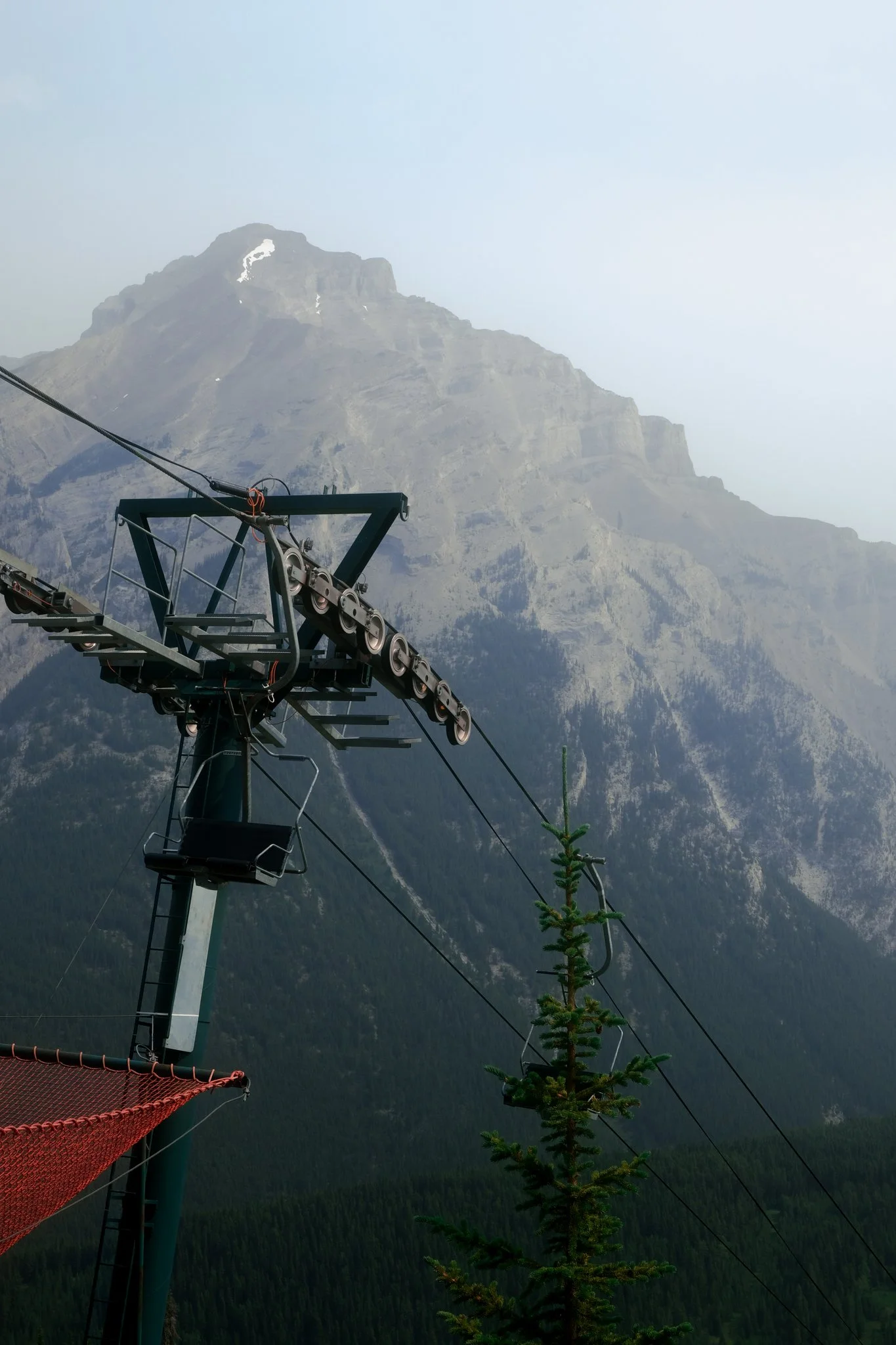 View of a mountain with a ski lift or chairlift in the foreground, marked by a tall pole, with a forested hillside at lower elevation and a cloudy sky overhead.