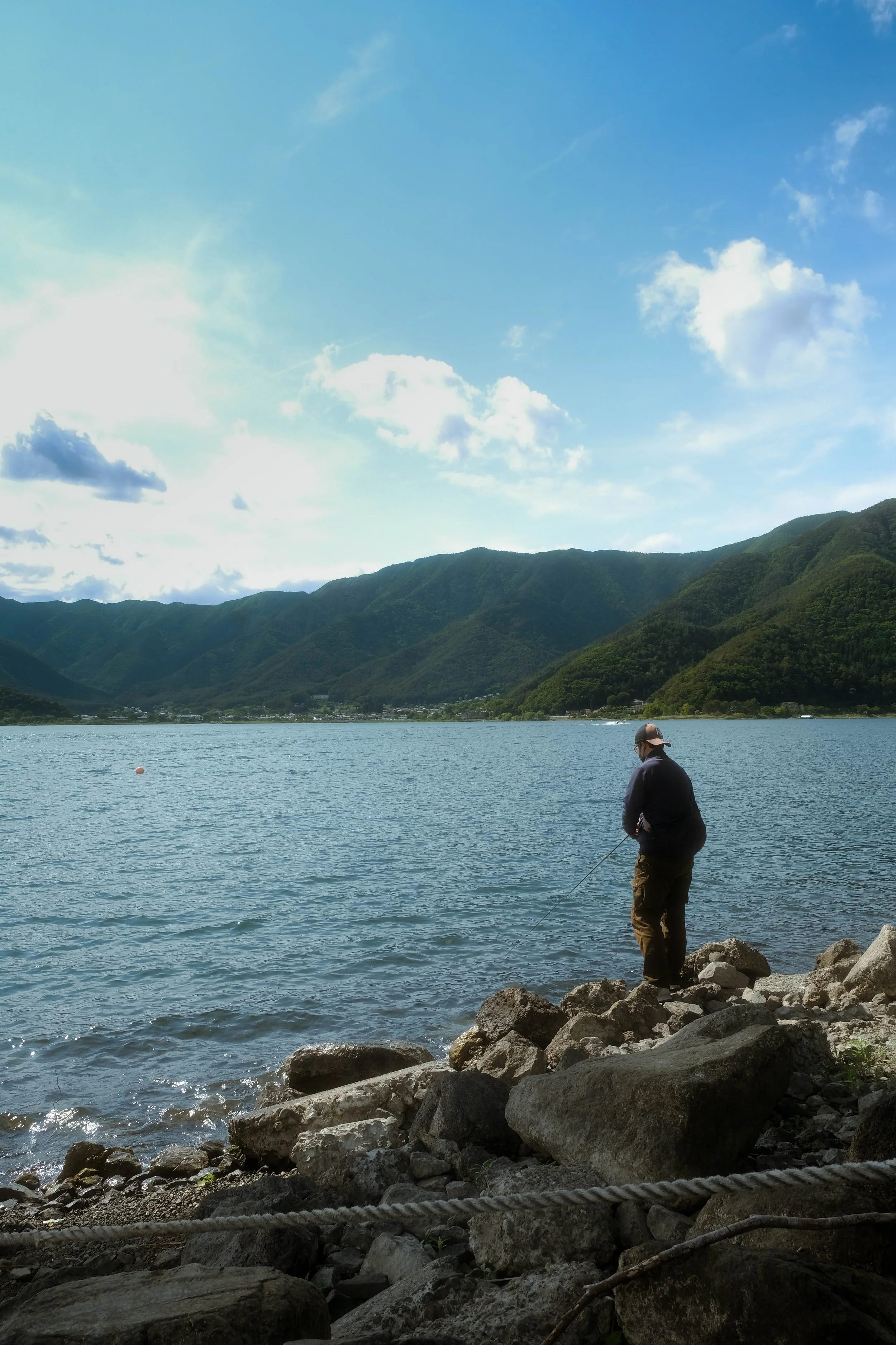 A person standing on rocks by a body of water, fishing, with mountains and a partly cloudy sky in the background.