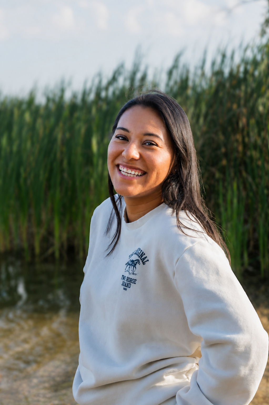 A young woman with shoulder-length dark hair and a bright smile stands outdoors near a body of water and tall green reeds, wearing a white sweatshirt with a logo on the chest.
