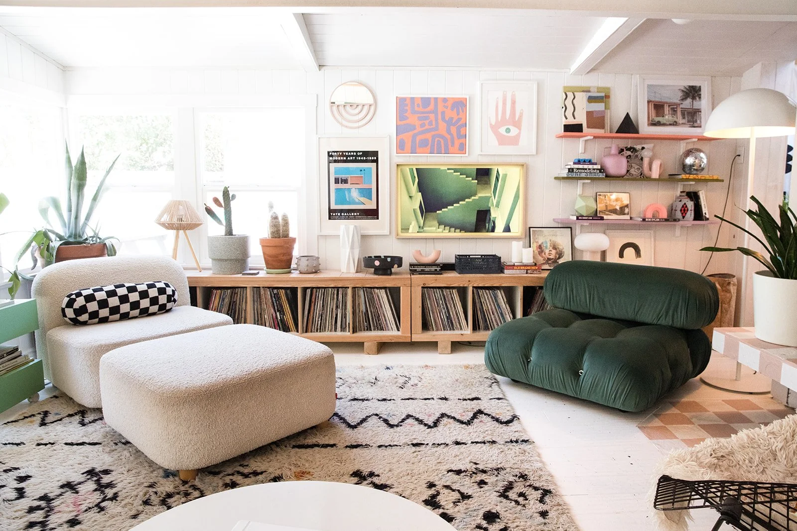 Living room with white walls, a white sofa with a checkered bolster pillow, a green lounge chair, a patterned rug, and a wooden media console filled with vinyl records. Decor includes houseplants, framed artwork, and shelves with decorative objects, with large windows in the background letting in natural light.