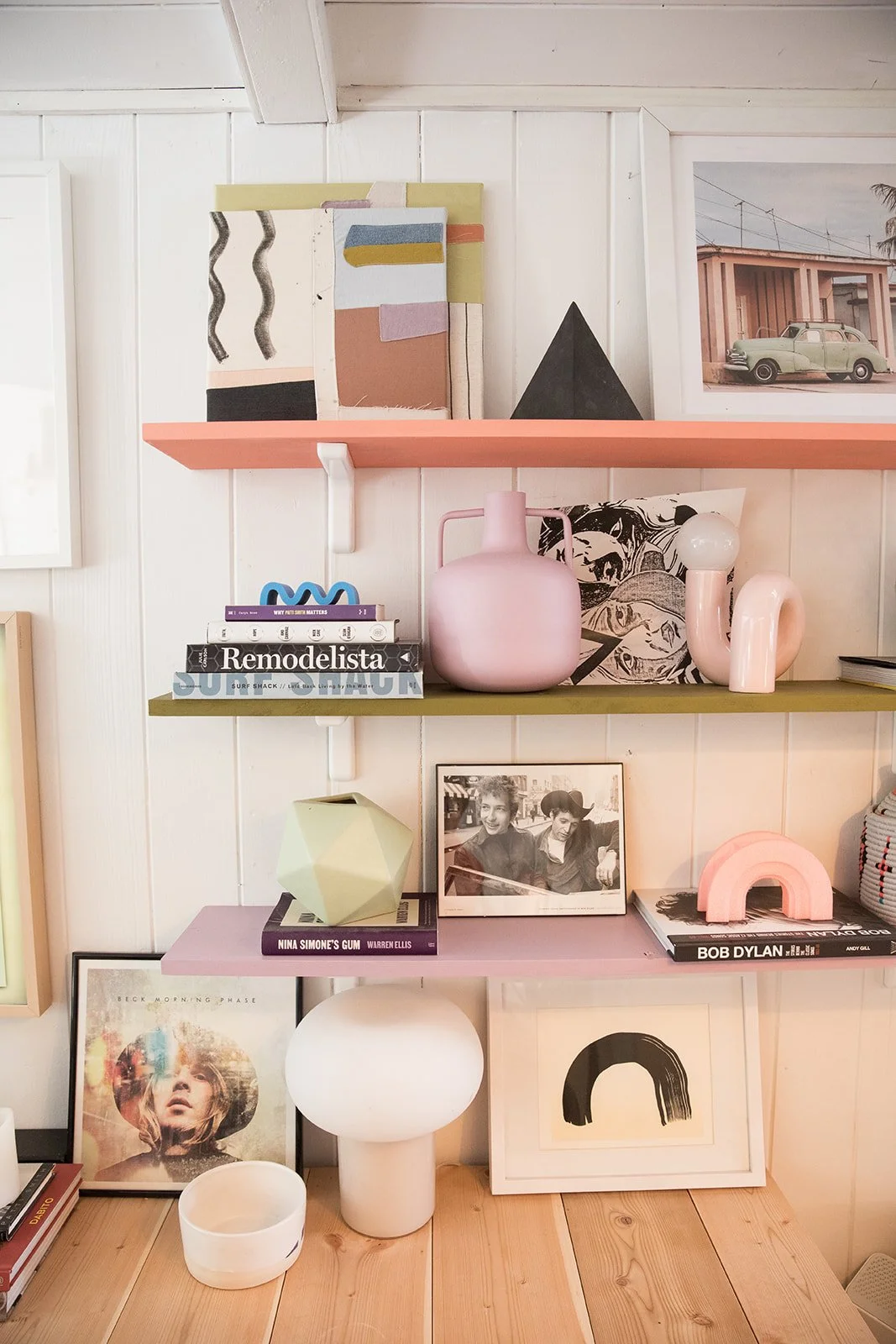 Decorative interior shelf with books, artwork, vases, and photographs. Items include pink and black ceramics, a framed drawing, and a vintage black and white photo of two people, set against a white paneled wall.