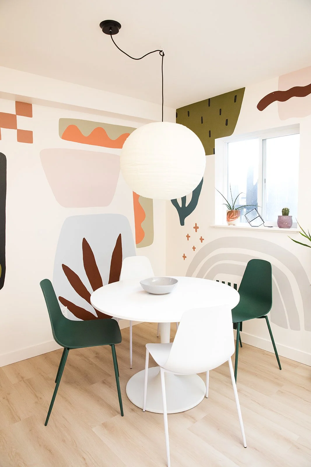 Modern dining room with a round white table and four chairs in white and green, abstract wall art mural, hanging paper lantern, window with small potted plants.