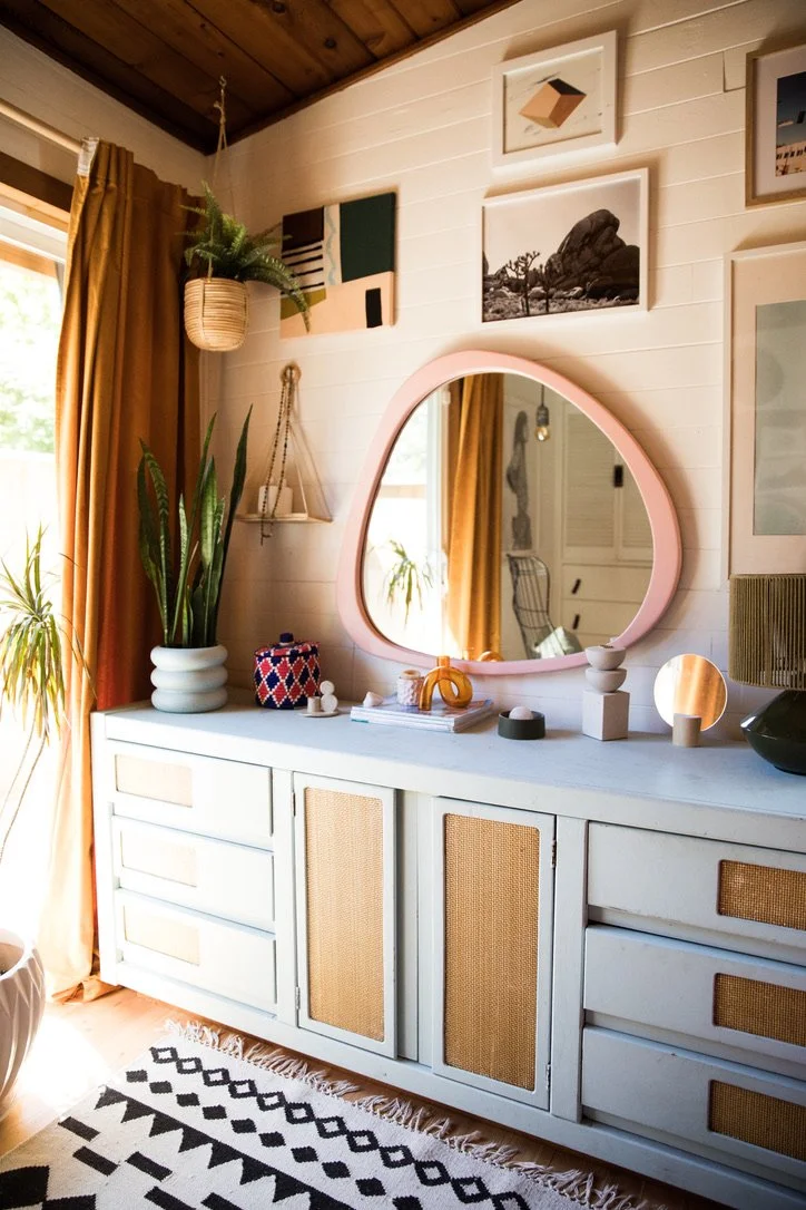 A white sideboard with rattan panel doors and drawers, topped with decorative items including plants, a pink mirror, and various small sculptures. Above the sideboard, several abstract and black-and-white photographs hang on a white wooden wall, with mustard-colored curtains and a window to the left. A patterned black and white rug is visible on the floor.