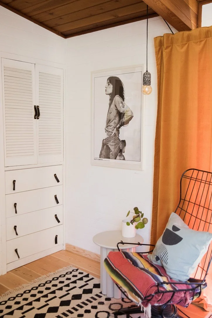 A cozy corner of a room with a black wire chair with a colorful blanket and a pillow. A small white side table with a potted plant, a mustard curtain, a framed black-and-white photo of a woman, and walls with wood paneling and white paint.