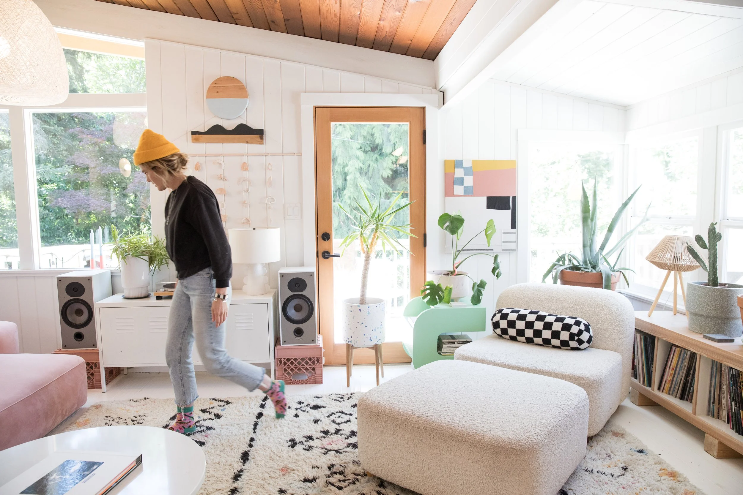 A woman with short hair, yellow beanie, black sweater, and gray sweatpants walking inside a bright, modern living room with white walls and wooden accents, surrounded by plants and furniture.