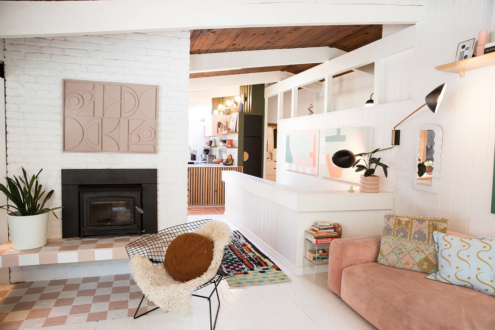 Living room with a white brick fireplace, potted plant, black fireplace insert, pastel pink and patterned pillows on a pink sofa, geometric patterned chair with a round cushion, colorful rugs, abstract wall art, decorative mirror, black wall-mounted lamp, and open kitchen with wooden accents and shelves in the background.