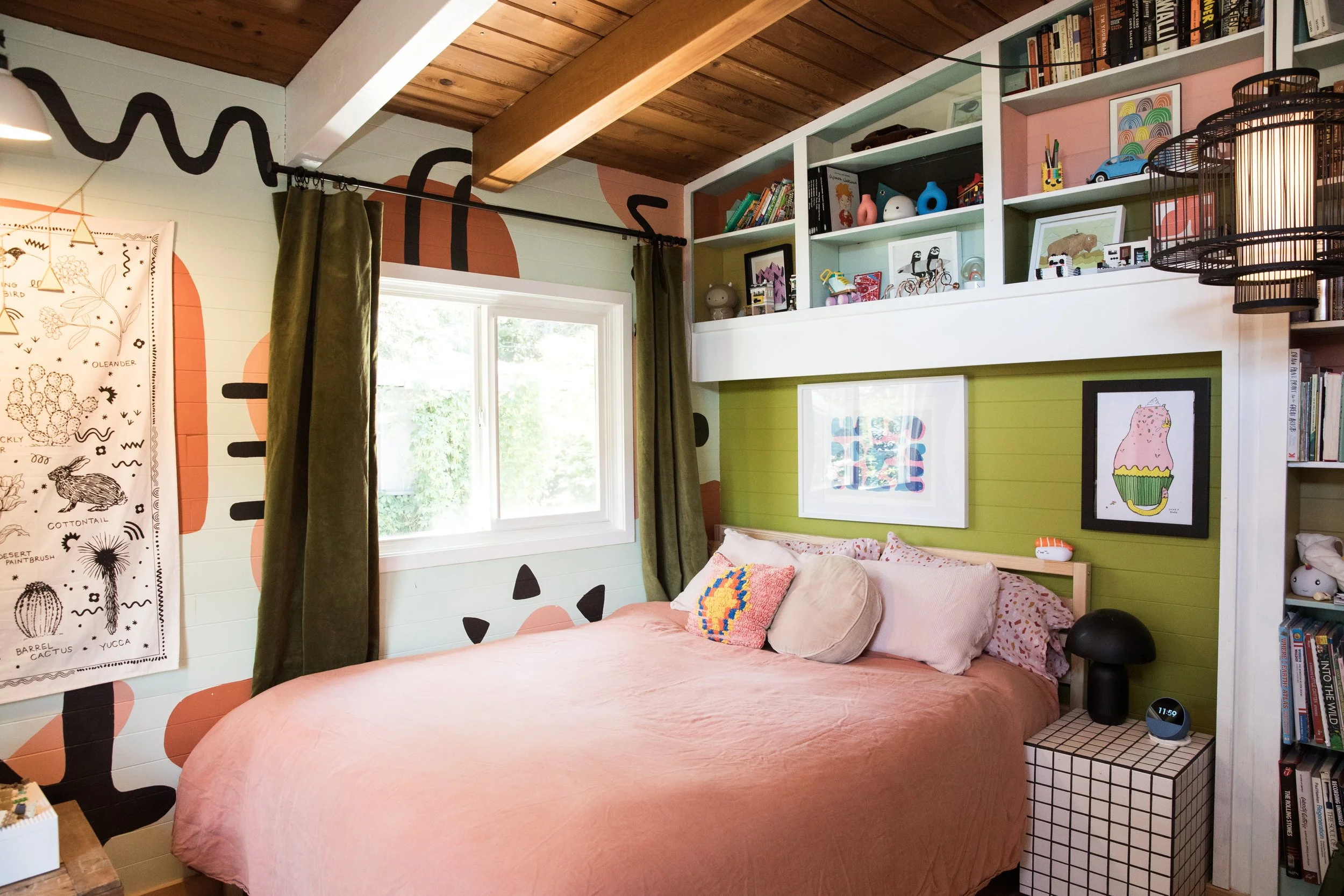 A cozy bedroom with a large bed covered in a peach-colored blanket, decorated with pink and colorful pillows. The room features a white built-in bookshelf filled with books, toys, and framed artwork, and has a piece of art with a pink cupcake on the wall behind the bed. There is a window with green curtains, and natural light streams in, revealing outside greenery. The ceiling is wooden with exposed beams, and part of a small black side table with a grid pattern is visible.