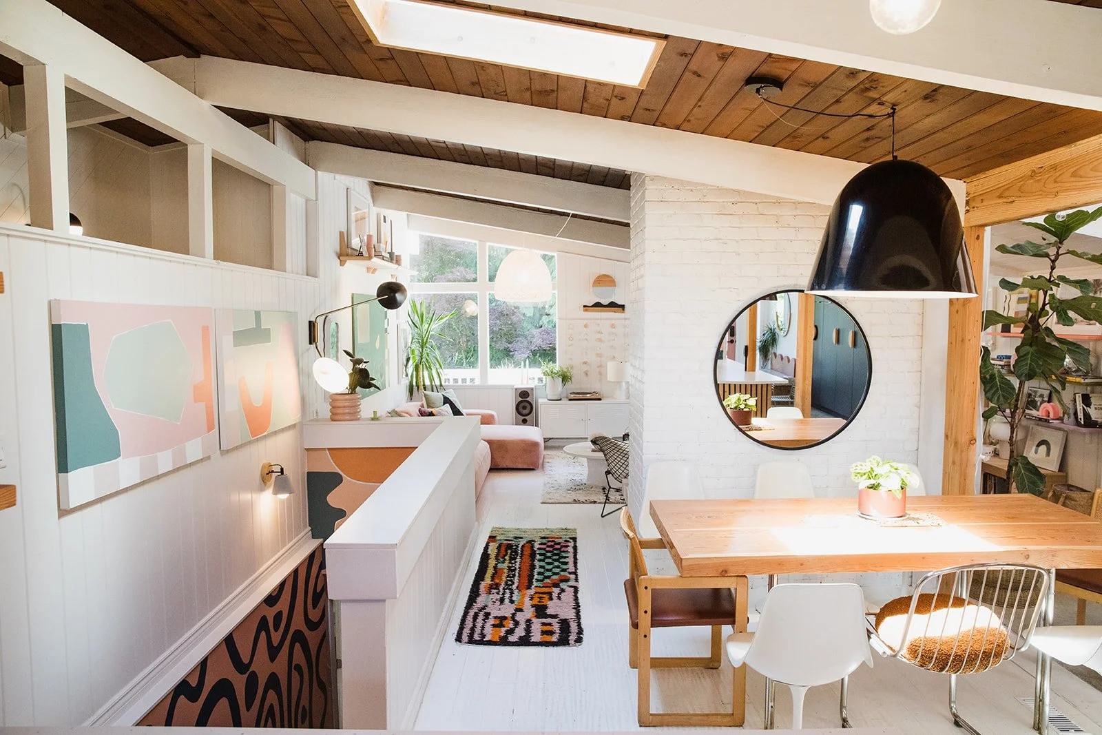 Bright, modern living room and dining area with natural light, white walls, wooden beams, and contemporary decor, including abstract art, plants, and round mirrors.