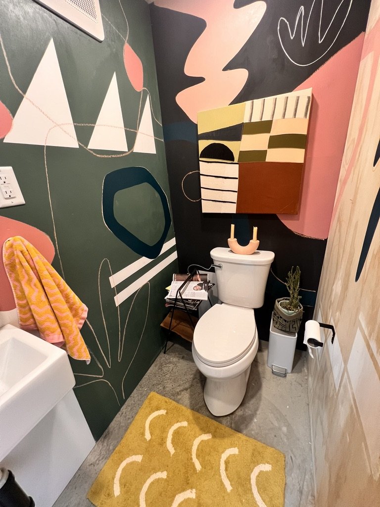 A small bathroom with abstract wall art, a yellow patterned rug, and colorful decor. Features include a toilet, small sink, a plant, a towel, and decorative items on a shelf.