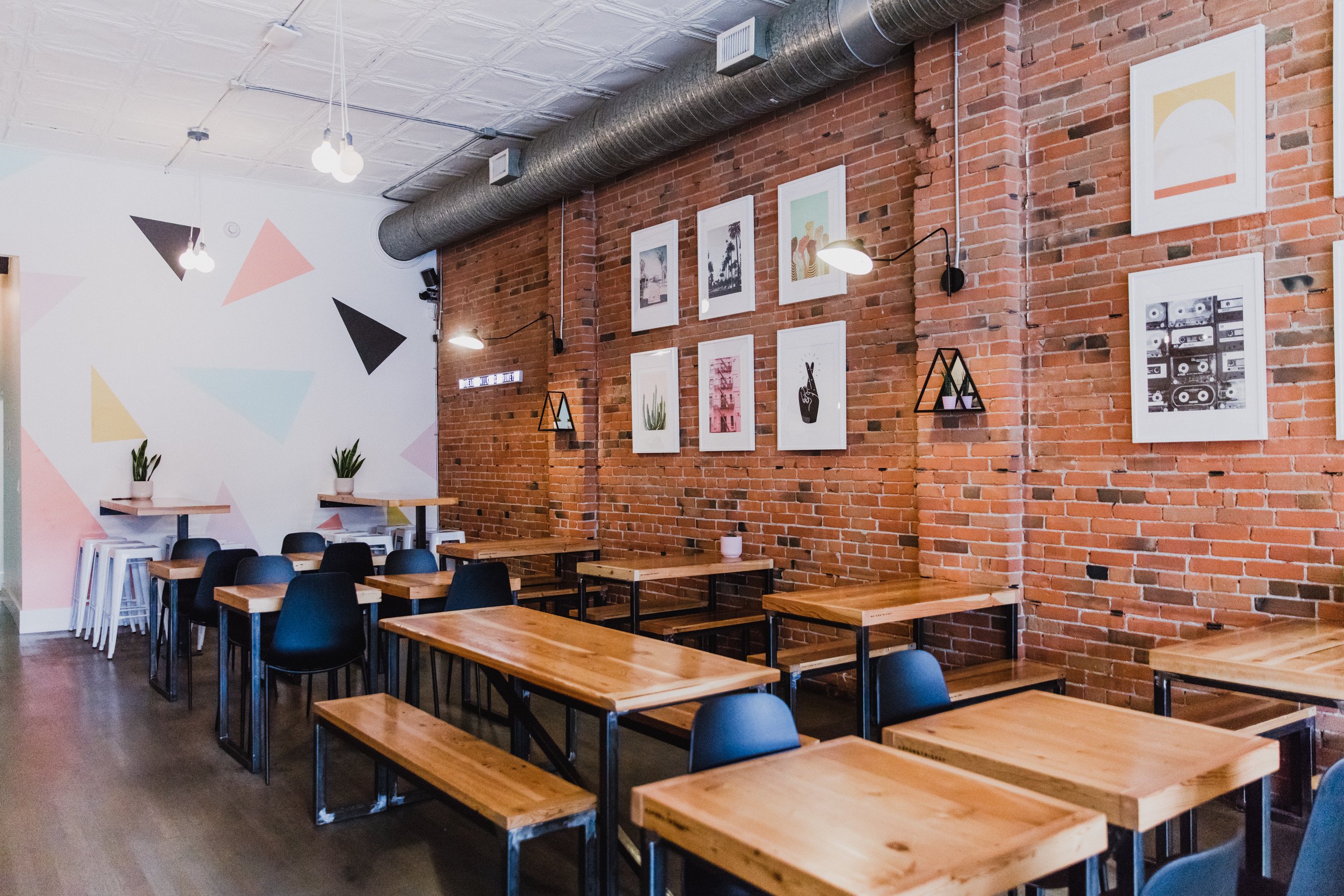 Interior of a modern cafe or restaurant with wooden tables and blue chairs, exposed brick walls decorated with framed artwork, potted plants, and industrial ceiling lighting.