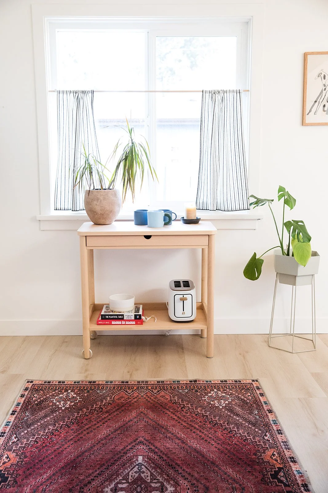 A minimalist living space with a window dressed in white and blue striped curtains, a small light wood console table with potted plant, mugs, candle, and books, a white countertop toaster, and a potted plant in a white stand next to a framed line drawing of a giraffe on the wall. There is a red patterned area rug on light wood flooring.