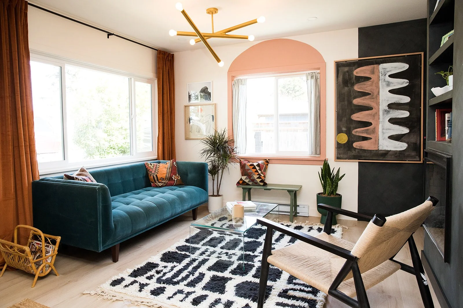 Living room with a teal sofa, abstract artwork, a large window with orange curtains, a glass coffee table, a patterned black and white rug, and a beige armchair.