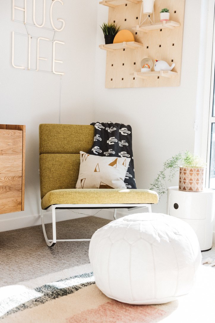A cozy living room corner with a green upholstered chair, a white cushion with geometric patterns, and a black and white blanket. There is a small white side table with a potted plant next to a window, and a white pouf ottoman in the foreground. To the left, part of a wooden cabinet is visible, and the wall behind features decorative wall art and a wooden pegboard with shelves holding plants and decorative objects.