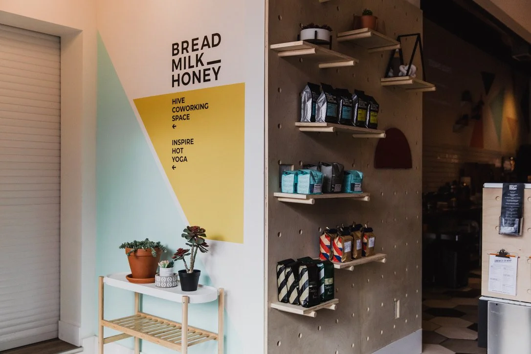 Coffee shop interior with shelves of coffee bags, a sign promoting a coworking space and yoga, and a small table with potted plants.
