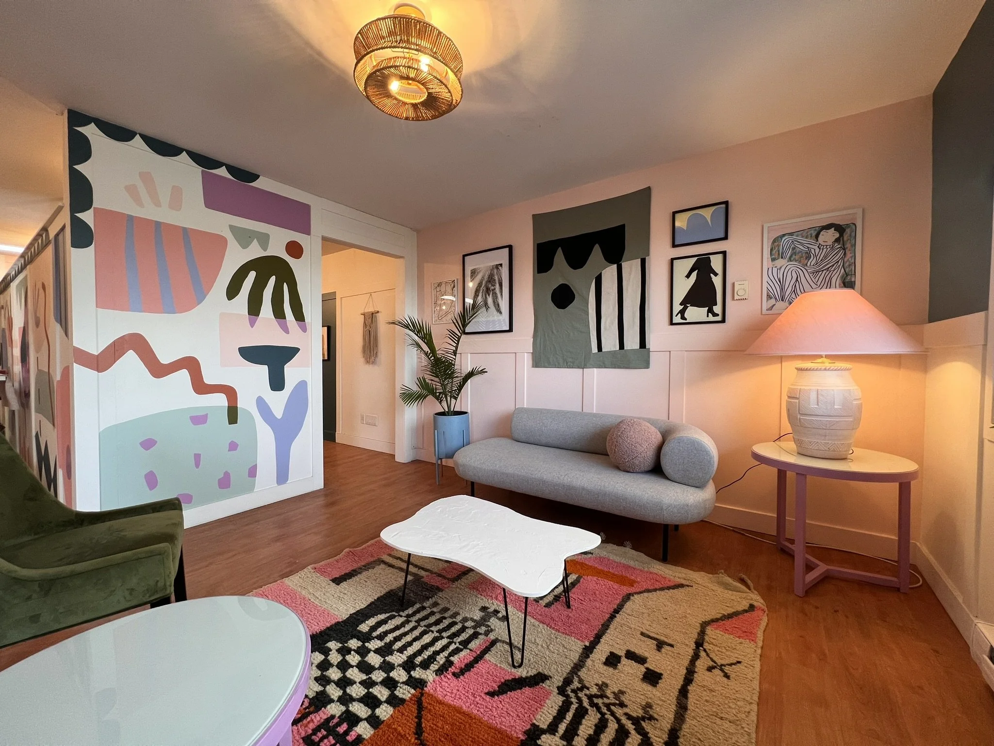 Living room with abstract artwork, framed pictures, a sofa, a small round table, a pink side table, a lamp, and a patterned rug.