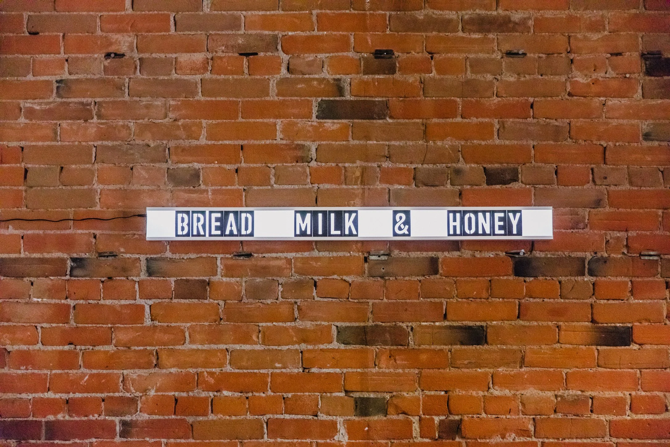 A brick wall with a lightbox sign that reads 'BREAD MILK & HONEY'. Modern wall art in a coffee shop.