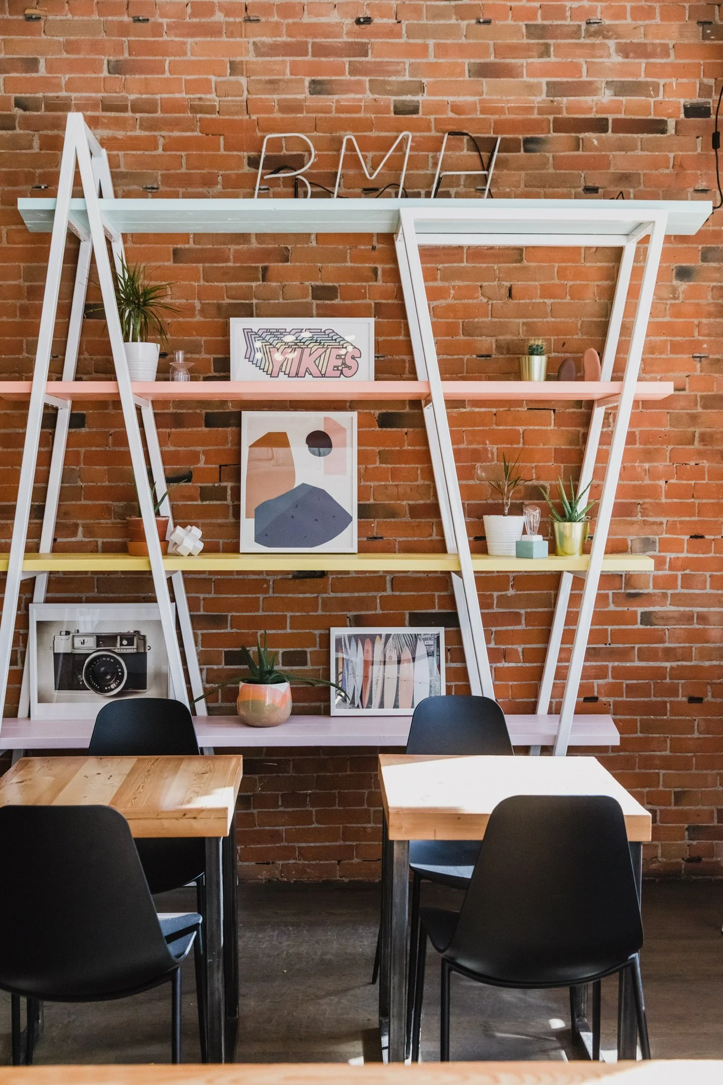 Interior of a modern cafe or coworking space with exposed brick wall, decorative plants, framed artwork, and black chairs around wooden tables.