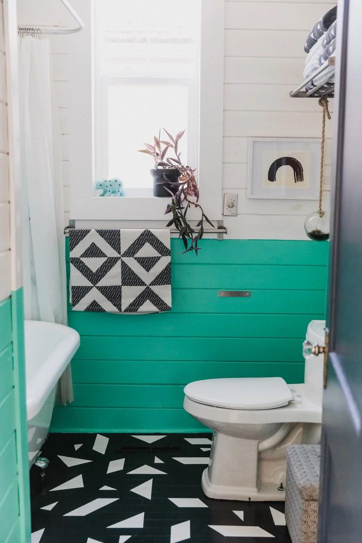 A small bathroom with white shiplap walls and a bright turquoise wainscoting. There's a white toilet, a black and white geometric patterned towel hanging on a rail, a small window with a plant and a decorative item on the sill, and modern black and white mosaic tile flooring. A framed abstract art piece hangs on the wall near the toilet.