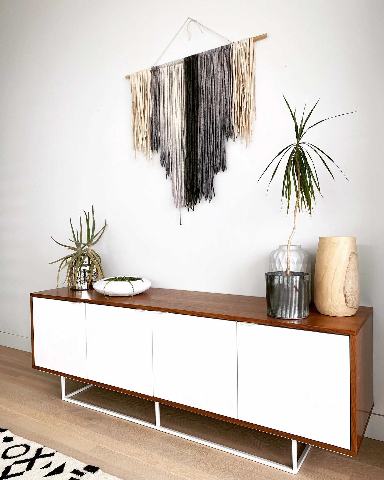 Modern wooden and white sideboard with decorative vases and potted plants on top, and a black and white rug in front.