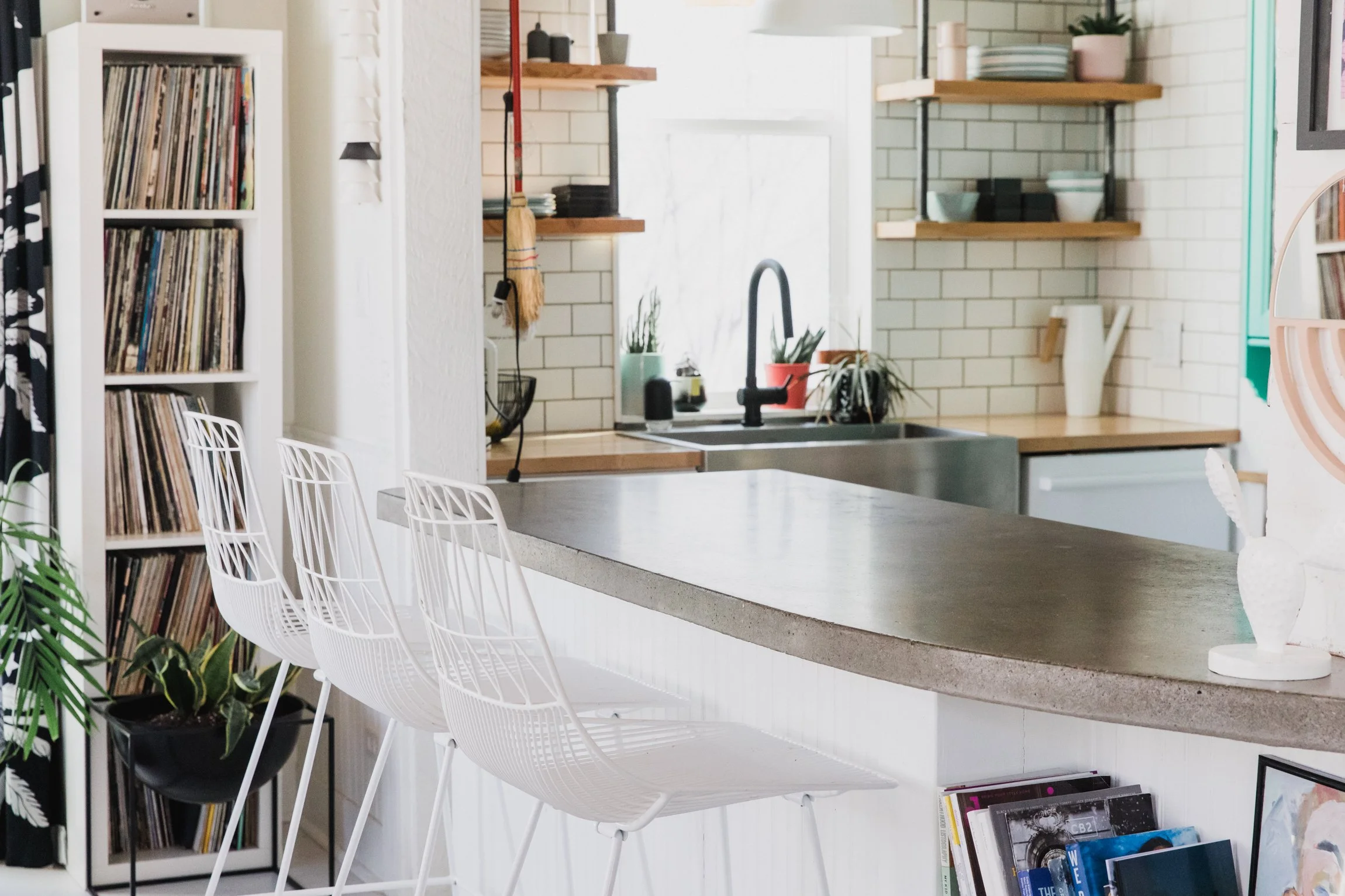 A modern kitchen with white cabinetry, open wooden shelves, a black faucet, a double sink, potted plants, and a kitchen island with white bar stools.