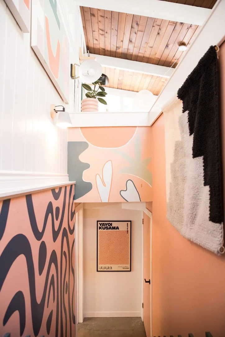 A narrow hallway with pastel-colored abstract wall art, a framed poster on the wall, and a large mirror reflecting a wooden ceiling with beams.
