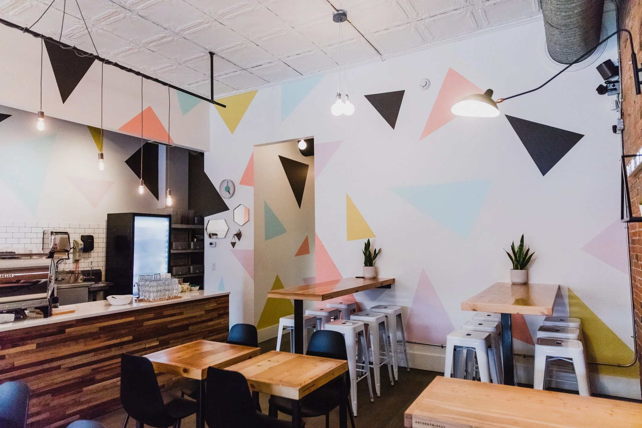 Interior of a modern cafe or restaurant with wooden tables, black and white chairs, a colorful geometric wall mural, hanging light bulbs, and potted plants.
