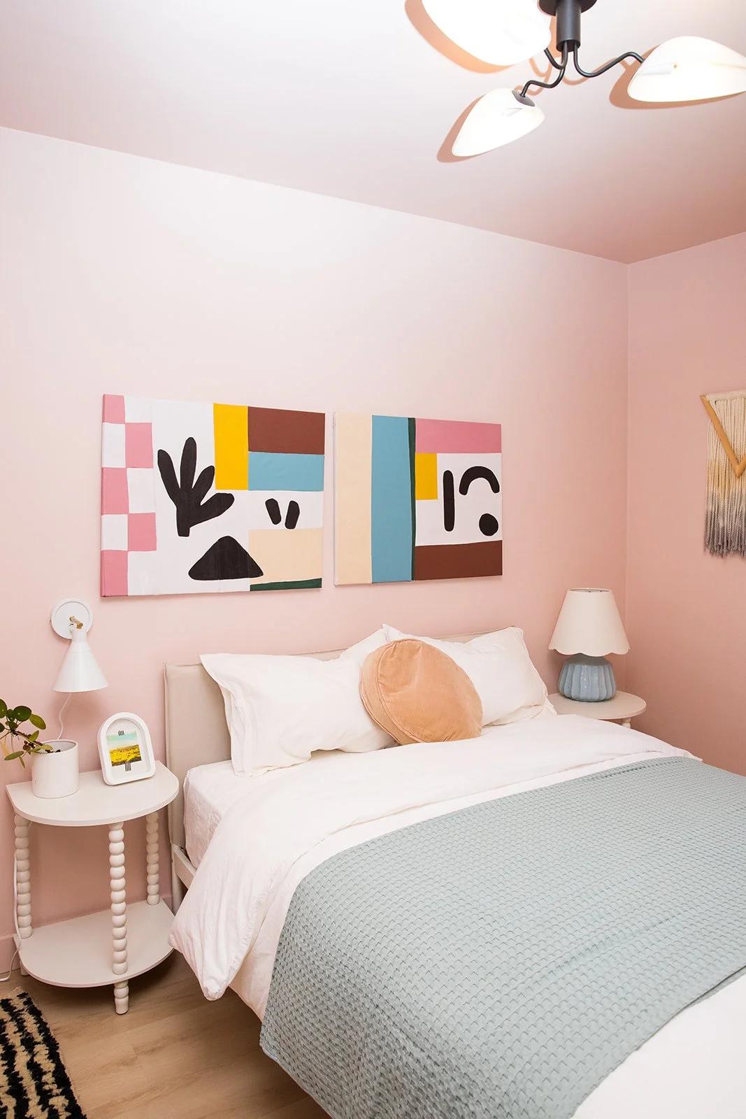 Bedroom with light pink walls, white bed with pillows and a mint green blanket, two abstract paintings on wall, nightstands with lamps, small plant and framed photo, ceiling light fixture.