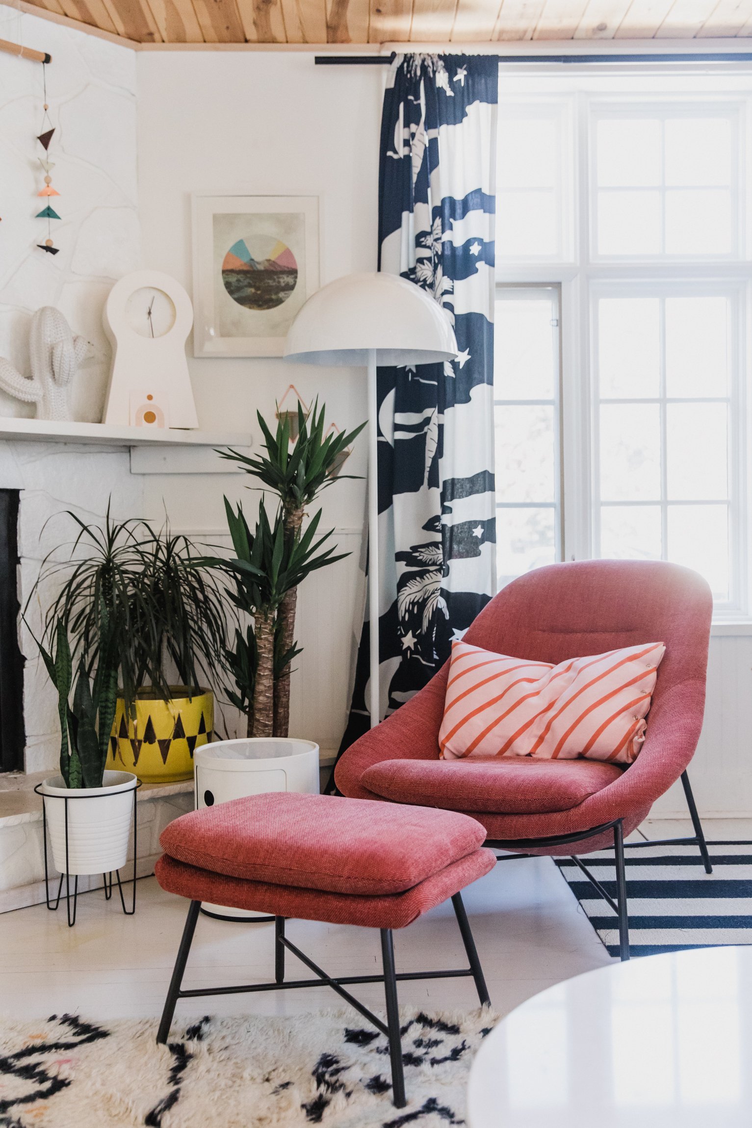 A cozy corner of a living room with pink armchair and ottoman, patterned throw pillow, potted plants in white and yellow planters, white side tables, and a white floor lamp. Light streams in from large windows with blue and white patterned curtains, a wall with desert-themed artwork, and a white mantle with decorative objects.
