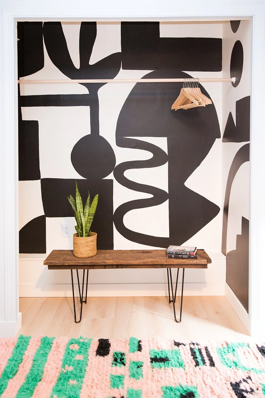 Room with a black and white abstract mural on the wall, a wooden bench with hairpin legs, a potted snake plant, a book, and a colorful pink and green patterned rug.