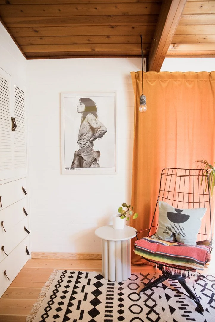 A cozy corner of a room with a white wall, wooden ceiling, and a black and white geometric rug. There is a white side table with a small potted plant, an orange curtain, and a black wire chair with a pillow and throw blanket. A framed black-and-white portrait of a woman hangs on the wall.