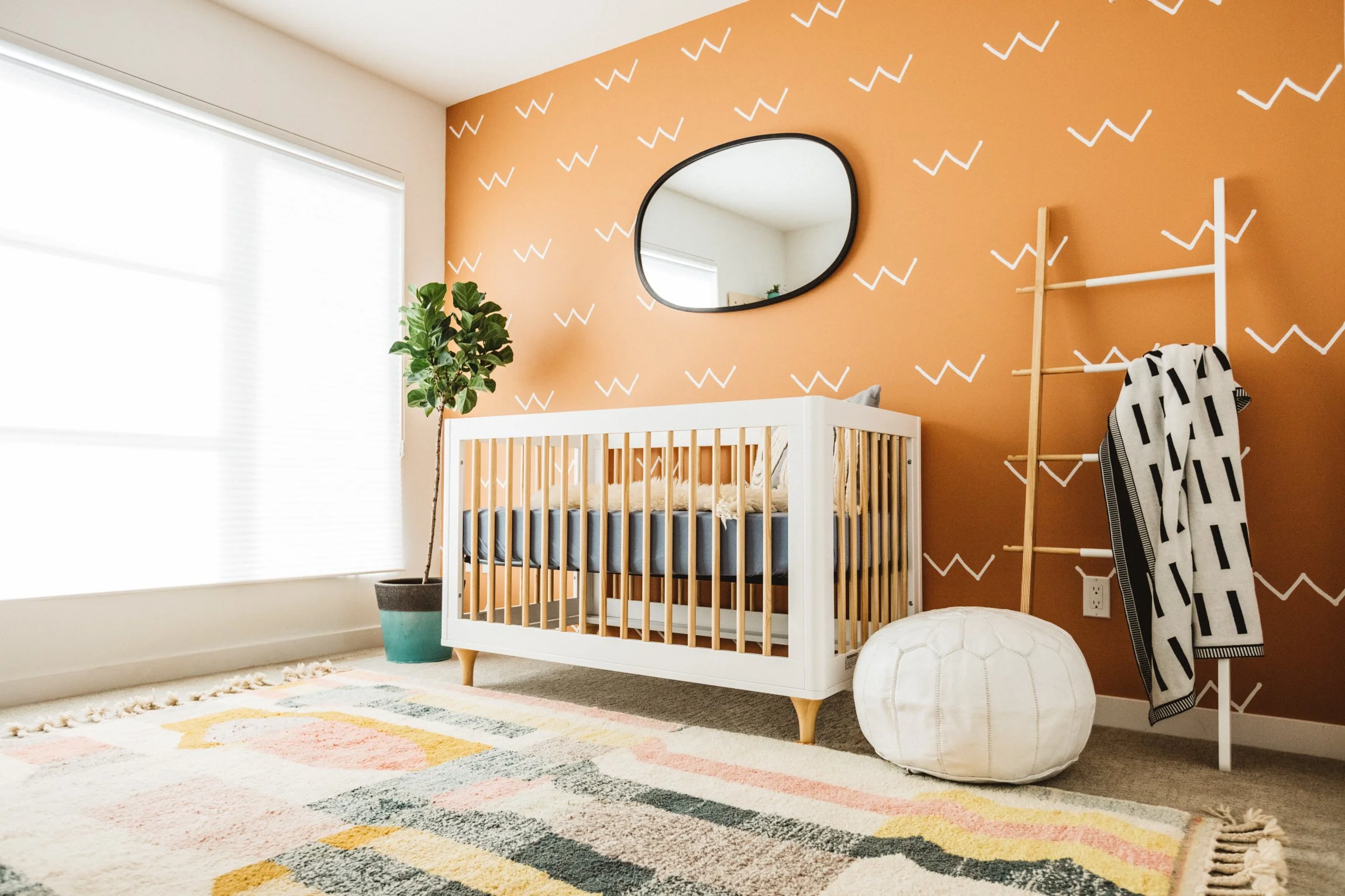 A nursery with rust tones and graphic pattern, a white modern crib. Featured in HGTV magazine. A potted fiddle leaf fig plant, a mirror with a black frame, a wooden ladder with a black and white blanket, a white pouf, and a colorful striped area rug.
