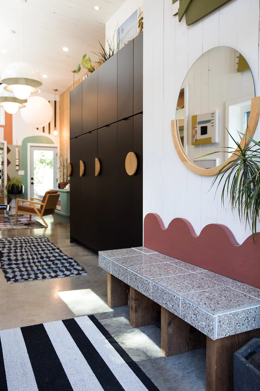 Interior of a modern, colorful home with a black cabinet, striped rugs, a mirror on a white wall, and a seating area with wooden chairs and patterned cushions.