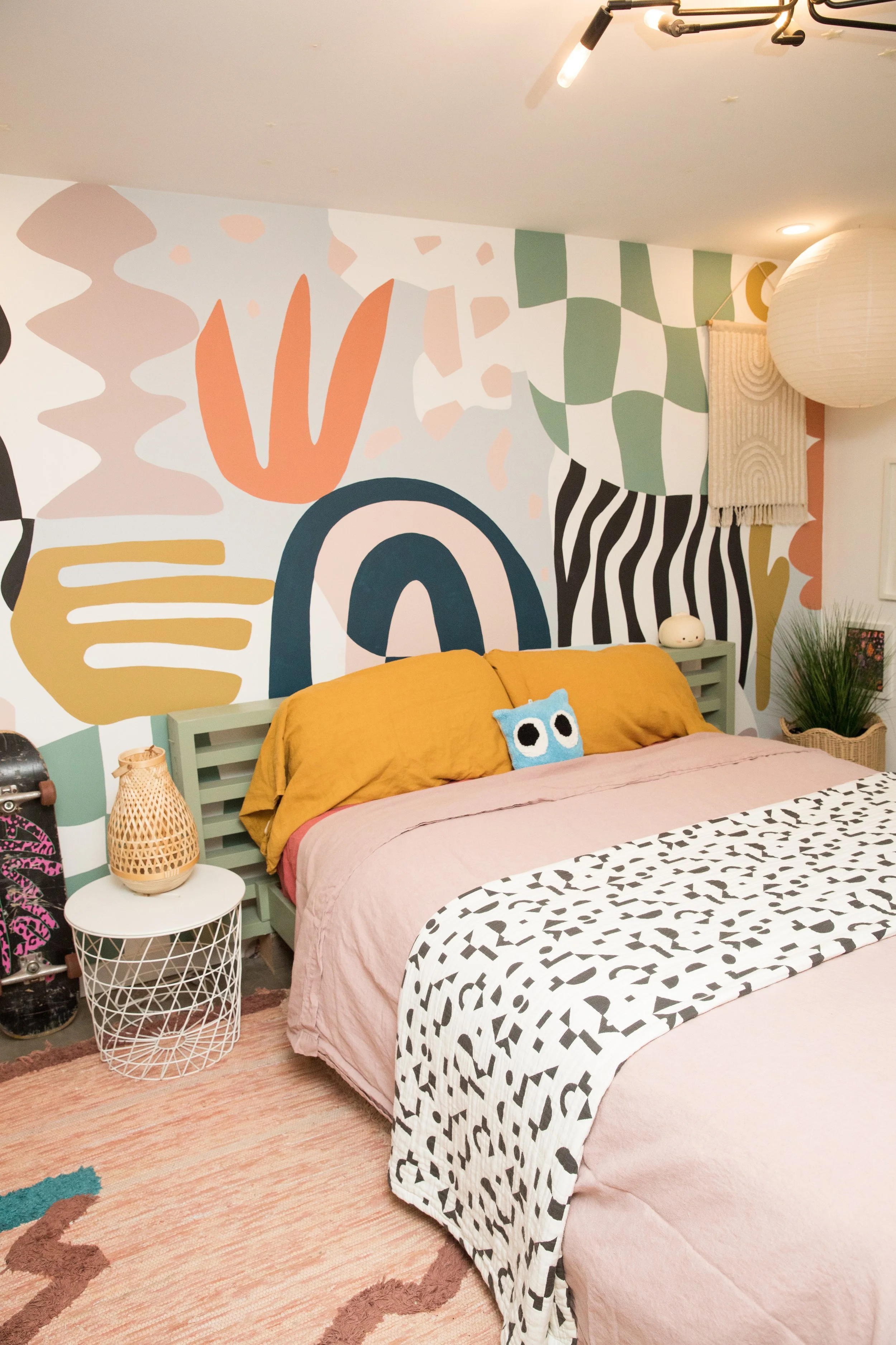 A bedroom with a mixed-pattern bedspread, a mustard-colored pillow, and a blue owl plushie. The wall features bold, abstract, colorful mural art. A white round side table with a woven lamp and a plant are beside the bed. The room has a wood floor and a pinkish rug.