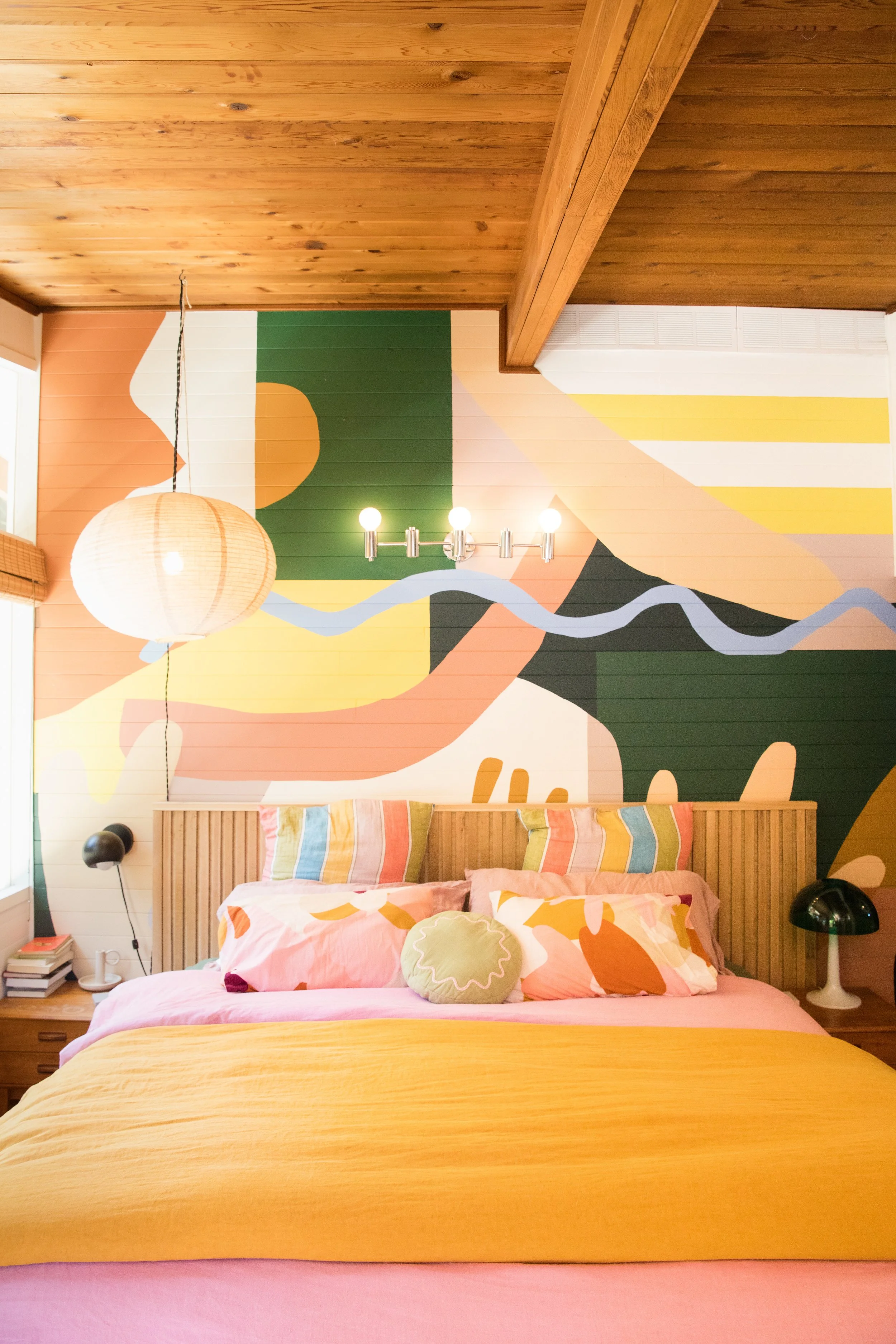 A bedroom with a wooden bed frame, colorful pillows, peach and yellow bedding, a large abstract mural on the wall behind the bed, two black bedside lamps, a paper lantern ceiling light, and a wooden ceiling.