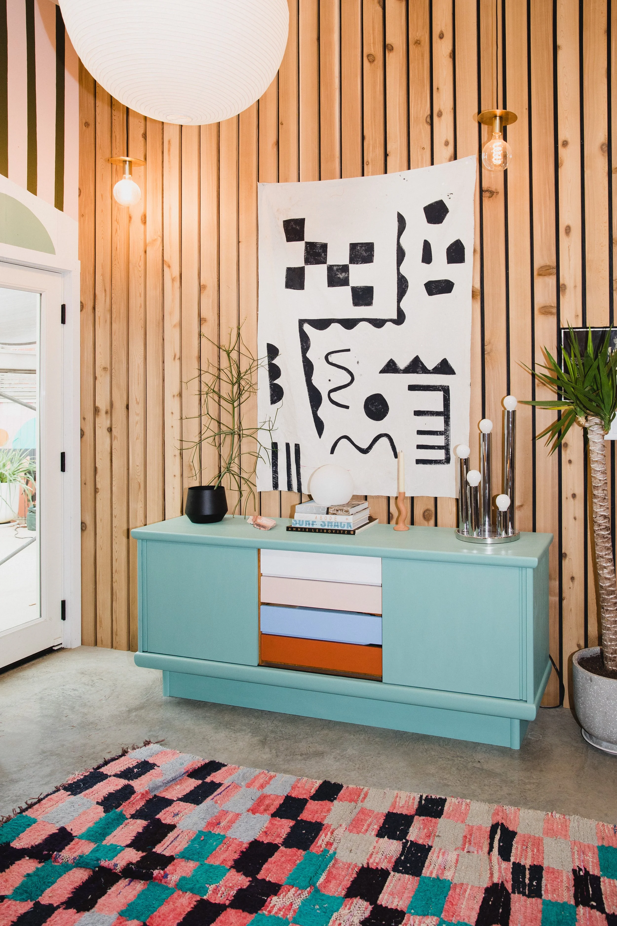 A turquoise sideboard with drawers and colored panels, decorated with a black vase and stacked books, standing against a wooden paneled wall with modern art, plants, and decorative objects, and a multicolored geometric rug on the floor.