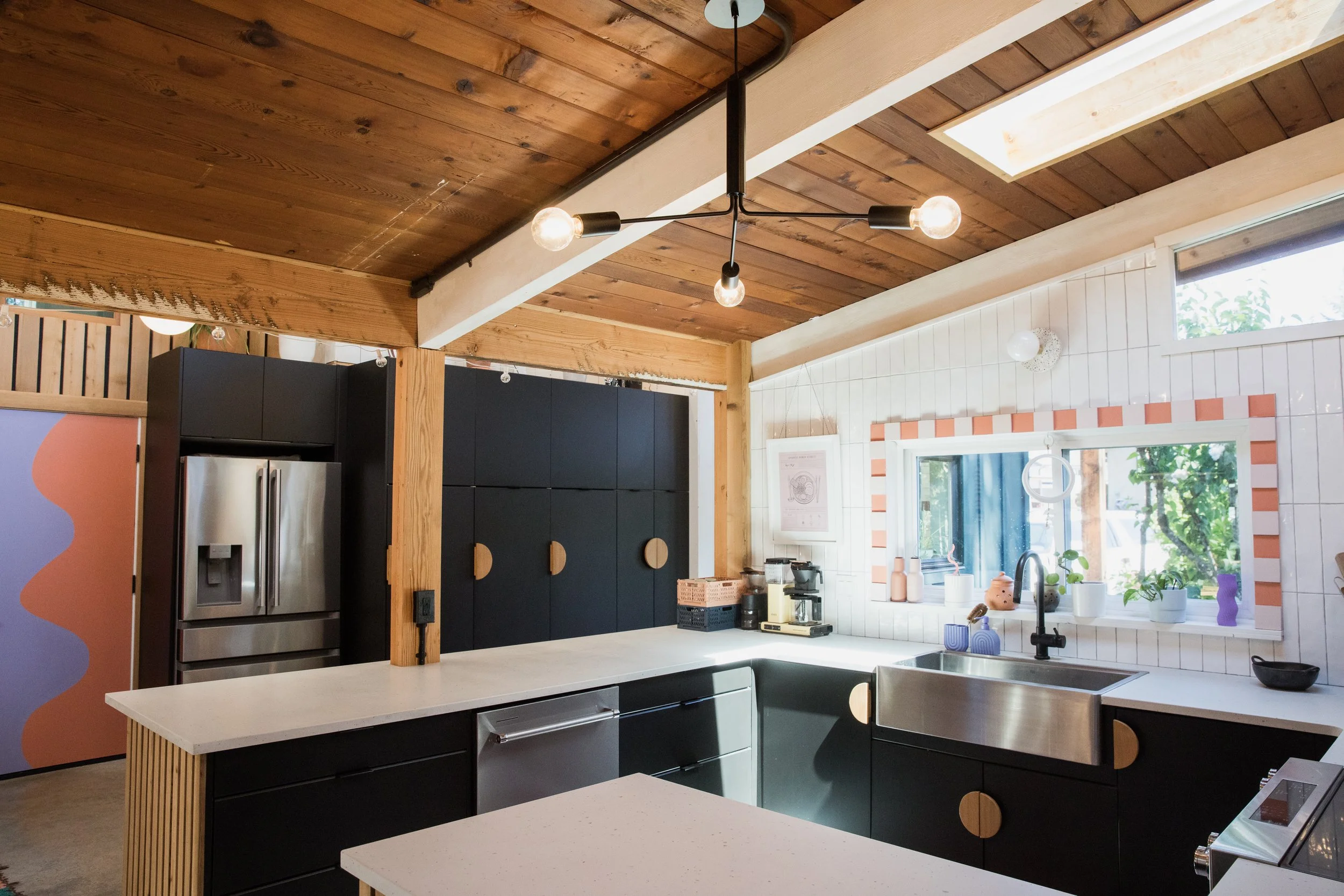 Modern kitchen with black cabinetry, white countertops, a stainless steel refrigerator, a black coffee maker, and various decorative plants on the window sill. The ceiling features wooden panels and a black minimalist chandelier with exposed bulbs.