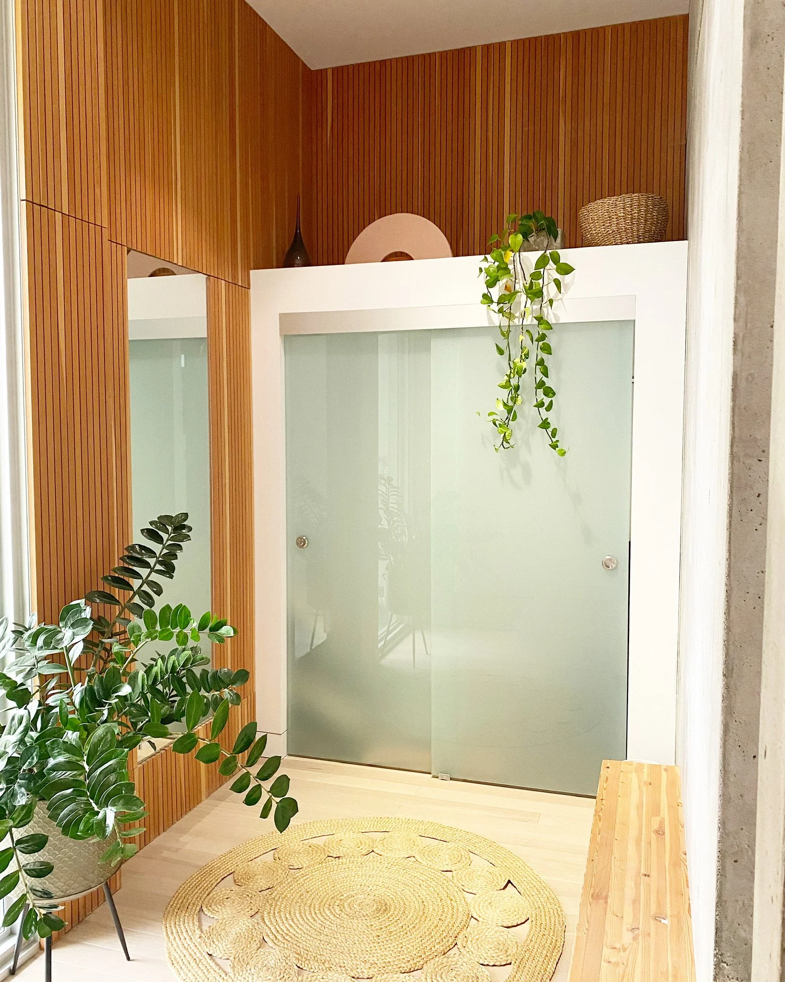 A modern interior space featuring a frosted glass door, wooden slatted wall, green plants, a woven rug, and a small wooden bench.